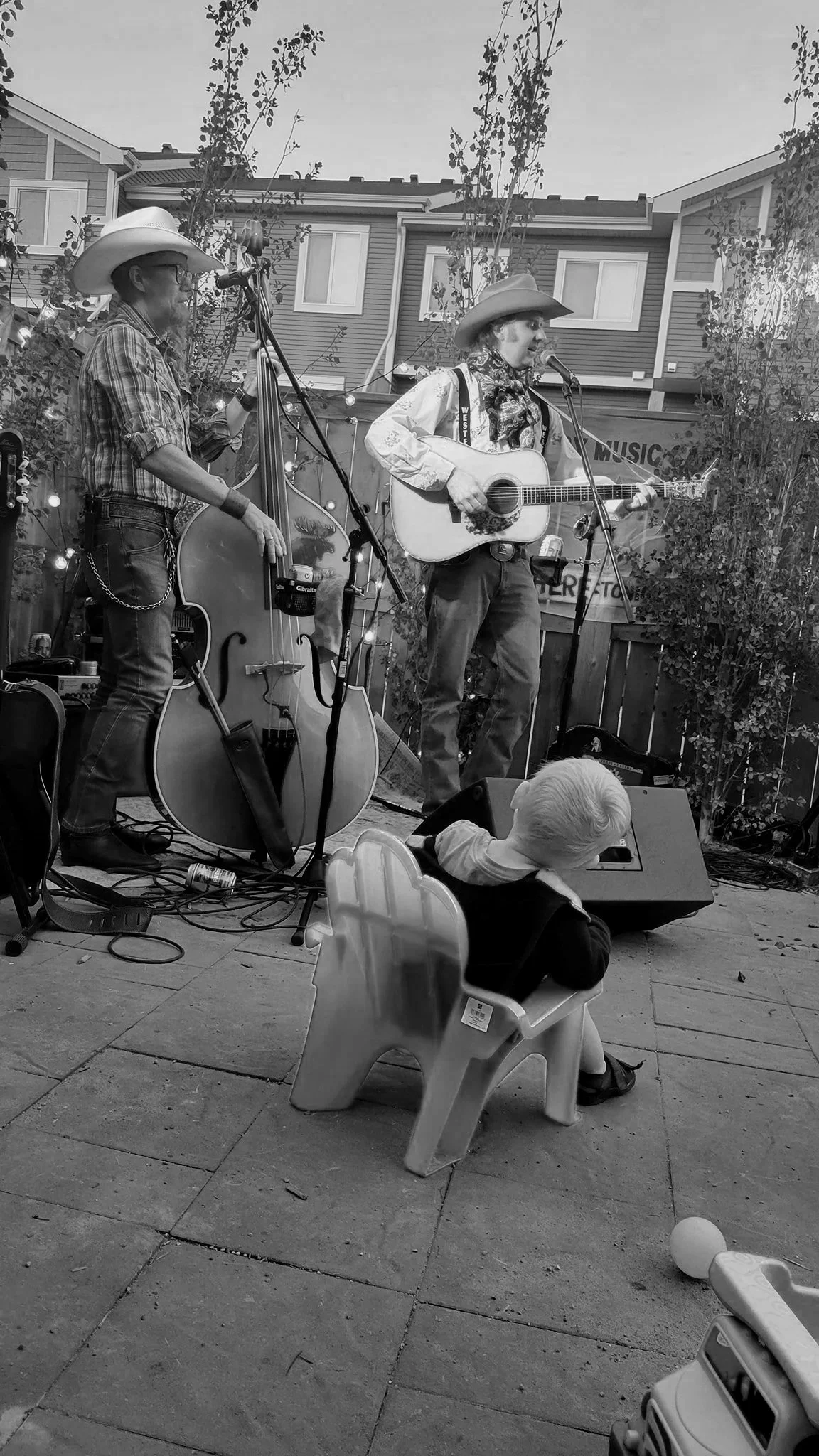 Tim Hus and his bassist Spider  perform outdoors: Spider is playing a double bass, and Tim Hus is singing and playing an acoustic guitar, both are wearing cowboy hats. A child is sitting in a small plastic chair watching in the foreground.