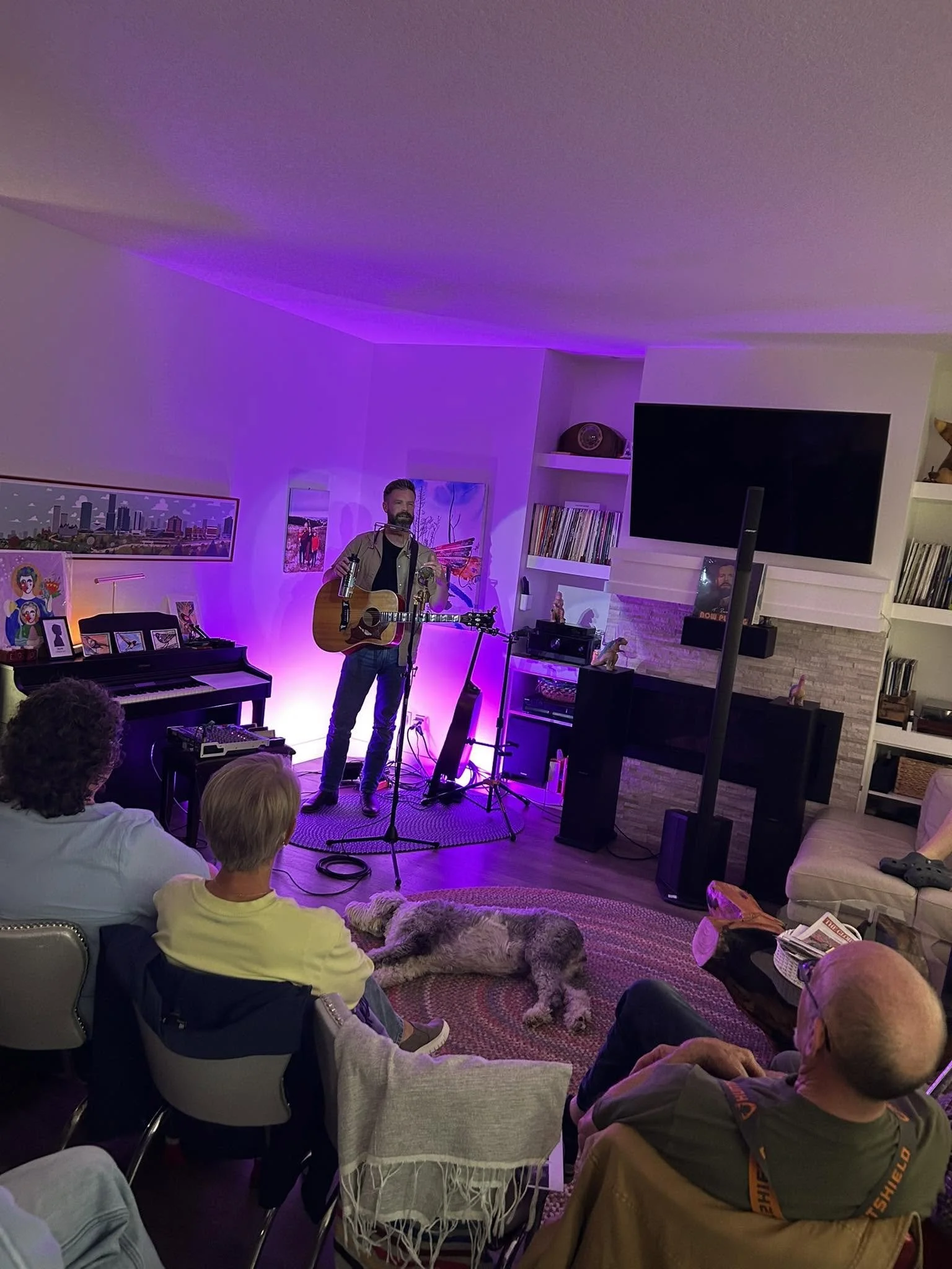 A man with a beard playing an acoustic guitar and singing into a microphone in a cozy living room with a TV, artwork, and bookshelves, engaging an audience seated on chairs and a dog lying on the carpet.