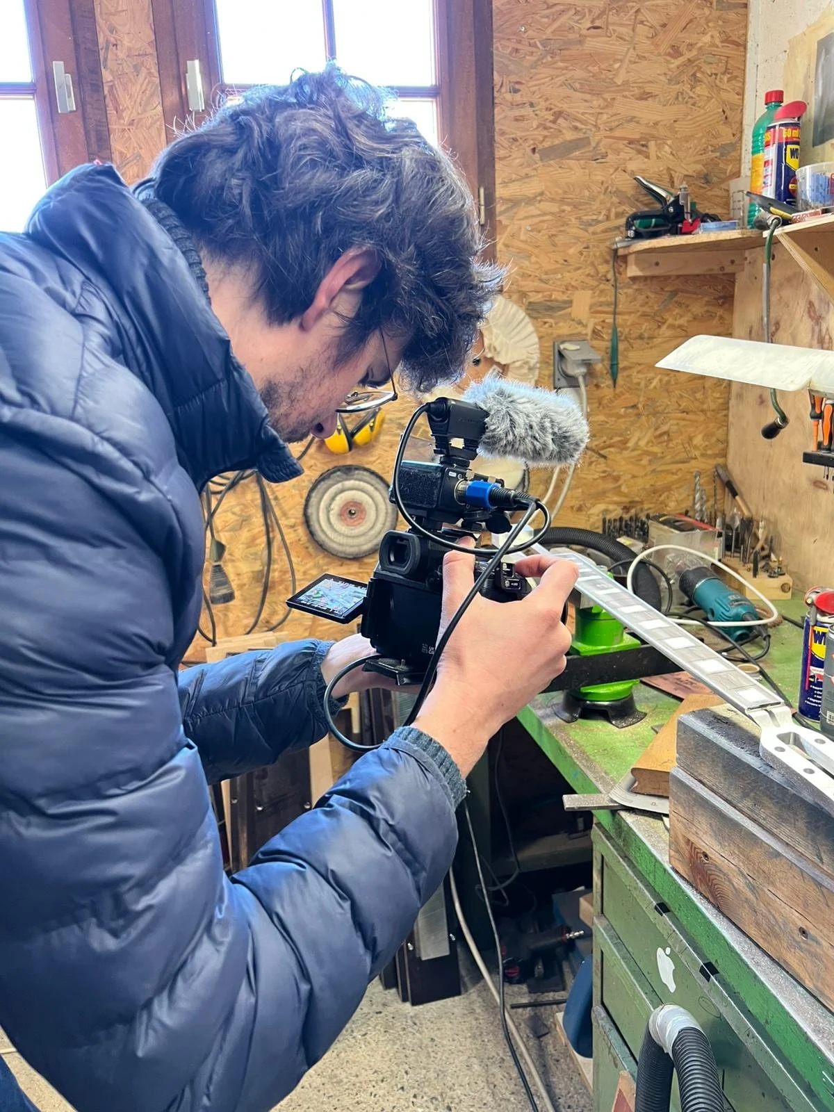 Un homme regarde dans un appareil photo dans un atelier, entouré d'outils et de matériaux de bricolage.