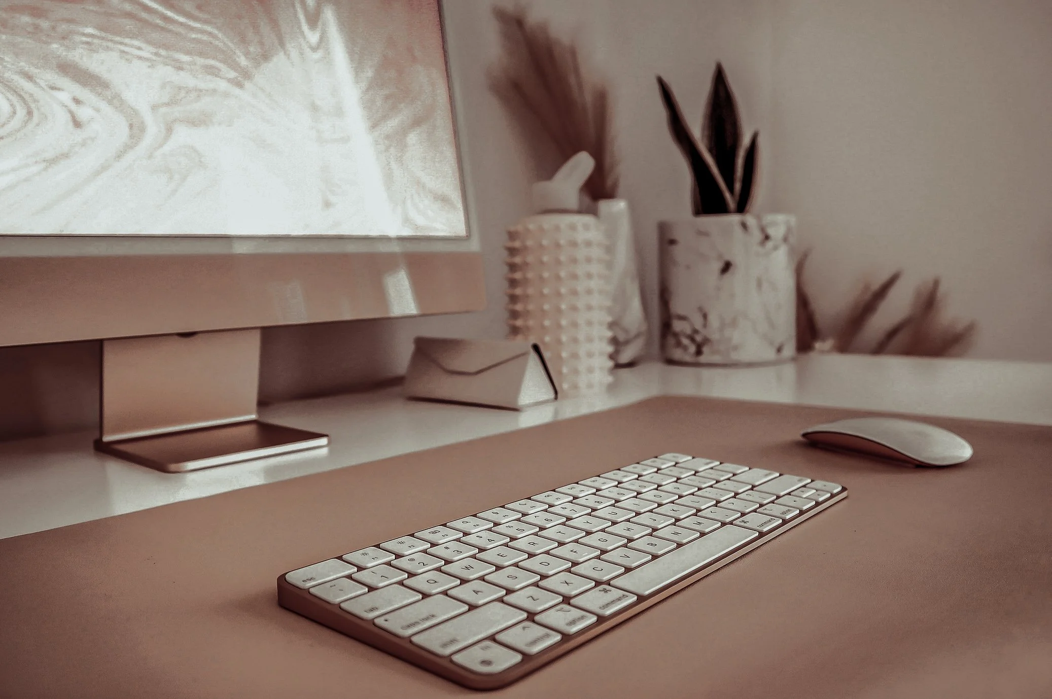 An Apple keyboard and Magic Mouse on a beige desk, with a computer monitor, a white lamp, and two vases with dried plants in the background.
