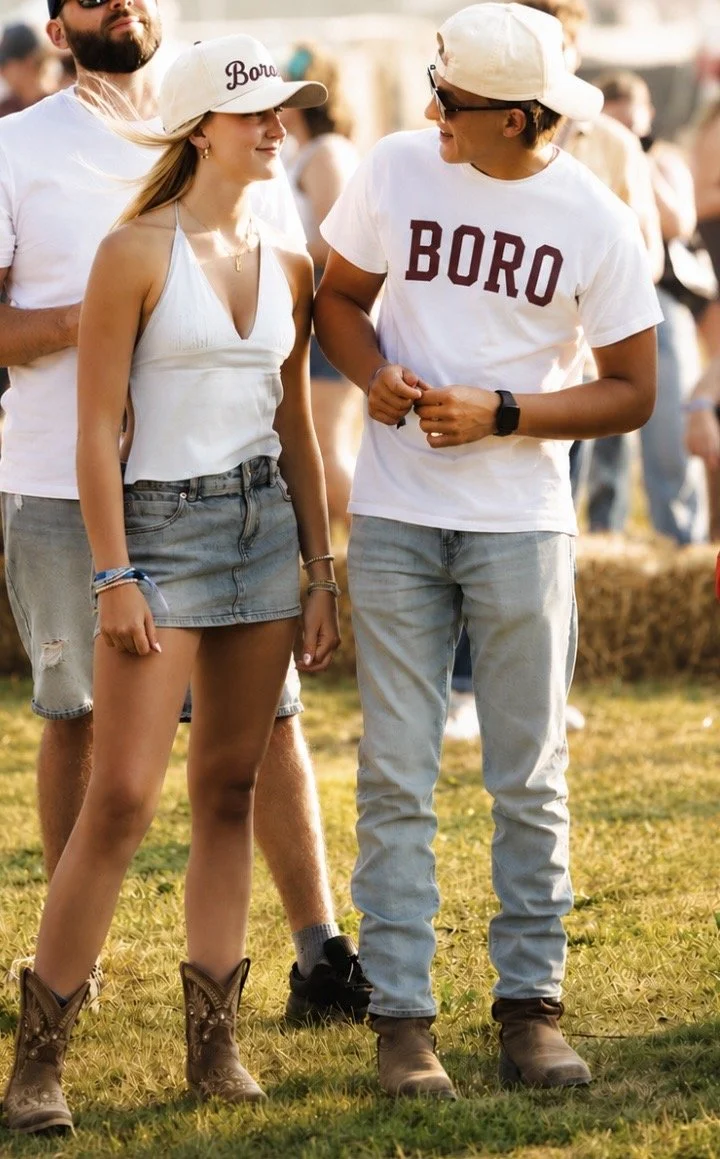 Two people, a woman and a man, are standing and talking outdoors at what appears to be a festival or fair. The woman is wearing a white tank top, a denim skirt, and cowboy boots, with a white cap and some jewelry. The man is wearing a white T-shirt with the word 'BORO' printed on it, light jeans, brown boots, sunglasses, and a white cap. There are other people and hay bales in the background.