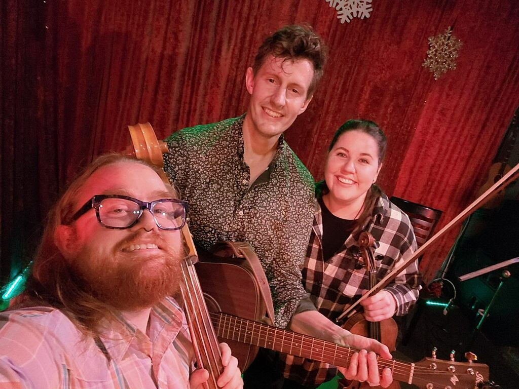 Three musicians smiling with musical instruments in a warmly lit room with red wooden walls and snowflake decorations.