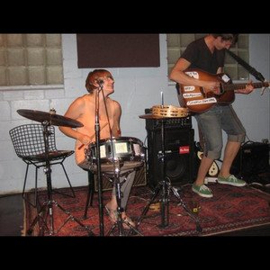 Two young men playing musical instruments in a recording studio. One is shirtless and seated at a drum set, while the other is standing and playing an electric guitar.