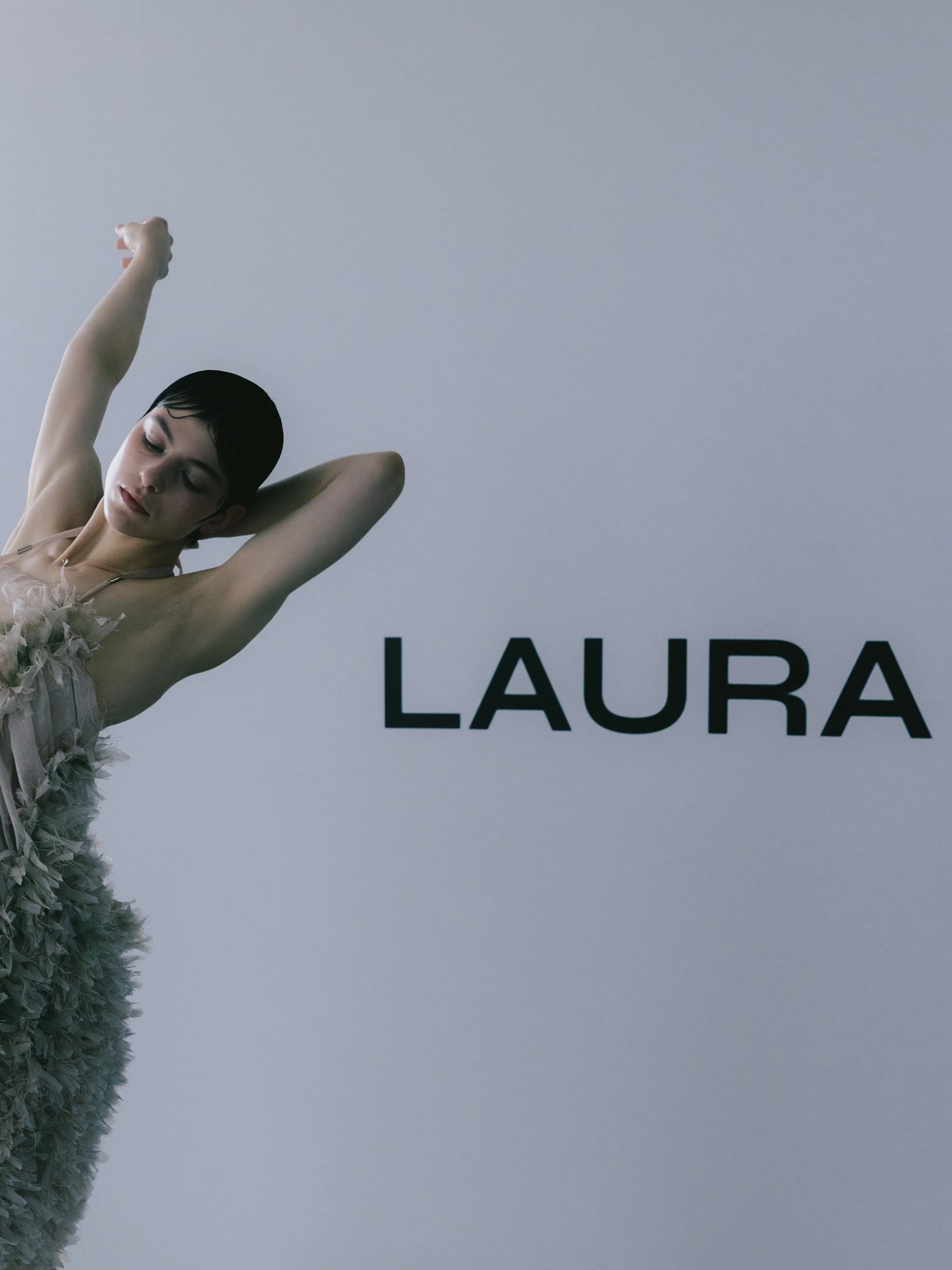 Ein junger Mann mit schwarzen Haaren, der eine elegante weiße Kleidung trägt, steht vor einem weißen Hintergrund mit schwarzen Buchstaben, die teilweise sichtbar sind, darunter der Name "LAURA".