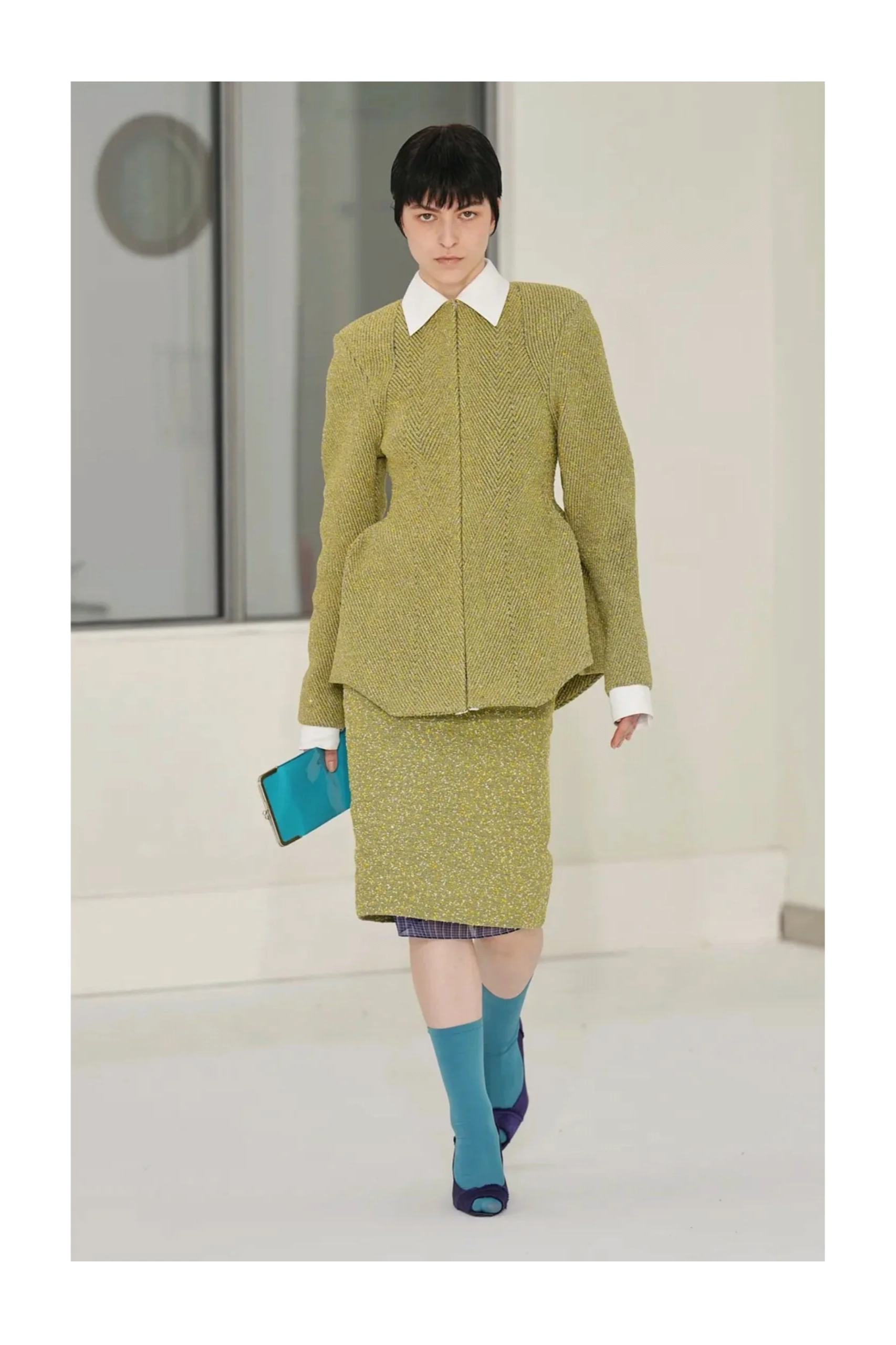 Ein Model läuft auf einem Laufsteg und trägt ein Outfit mit gelbem Oberteil, grüner Federboa, gelber Rock und blauen Stiefeln. Das Model hält ein Handy in der Hand.