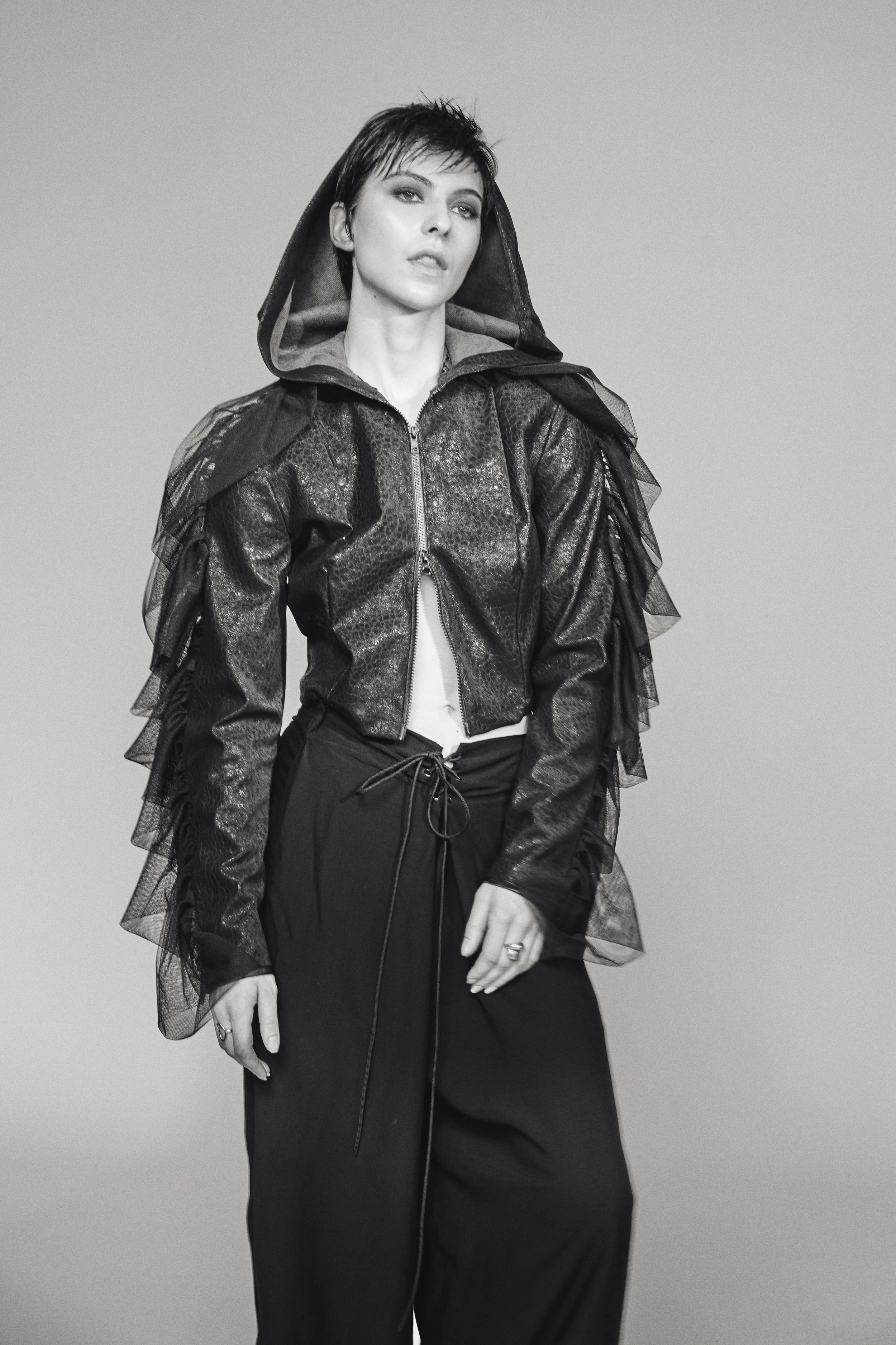 Schwarz-weiße Fotografie einer Frau mit kurzem schwarzen Haar, die in einem schwarzen Outfit mit einer Jacke mit Leder-Optik und Tüll-Details posiert