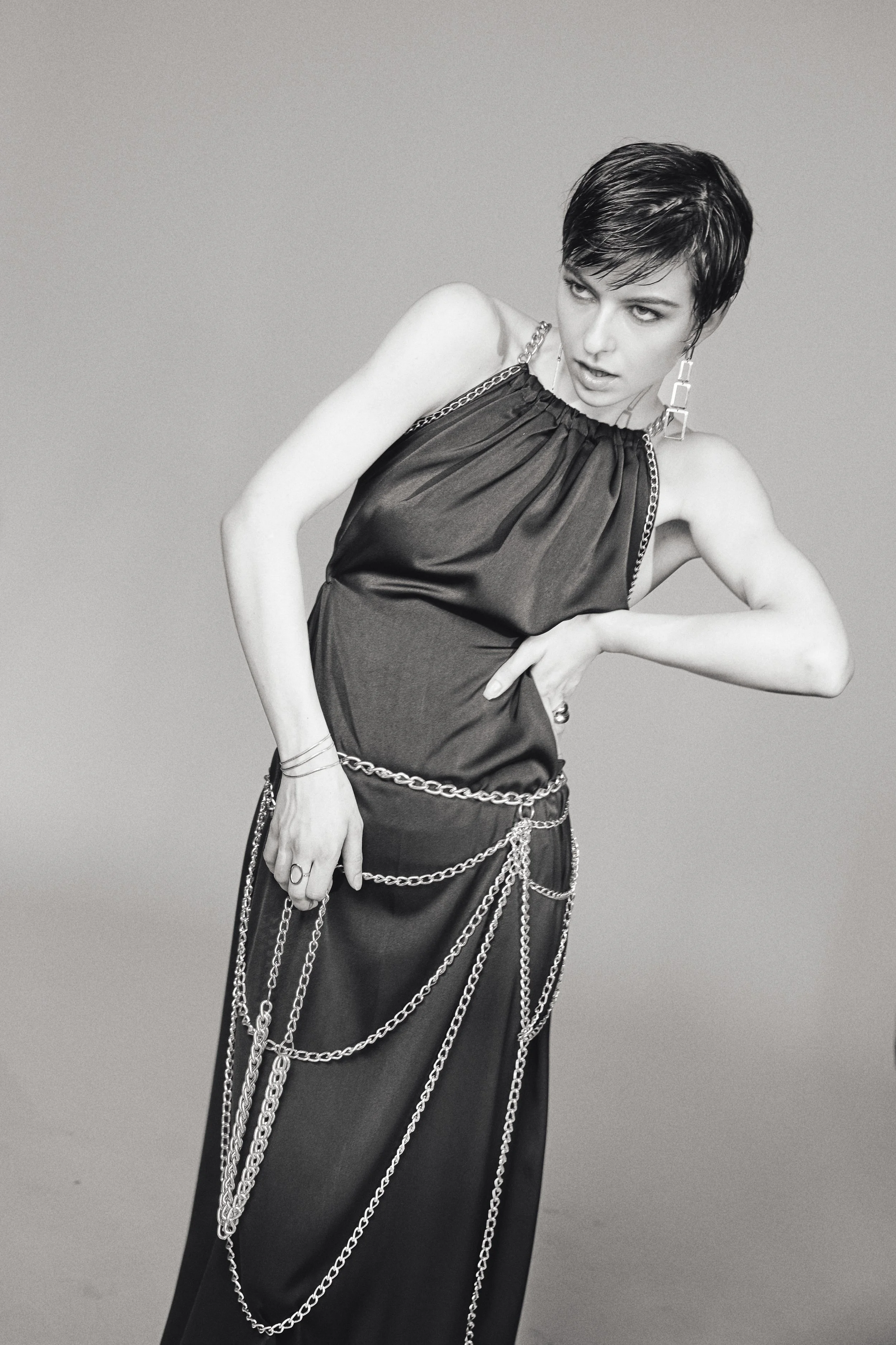 Schwarz-weiß-Foto einer jungen Frau mit kurzen, dunklen Haaren, trägt eine dunkle, glatte, elegante Abendkleidung und mehrere silberne Ketten sowie große Ohrringe, posiert mit einer Hand auf der Hüfte, gegen einen neutralen grauen Hintergrund.