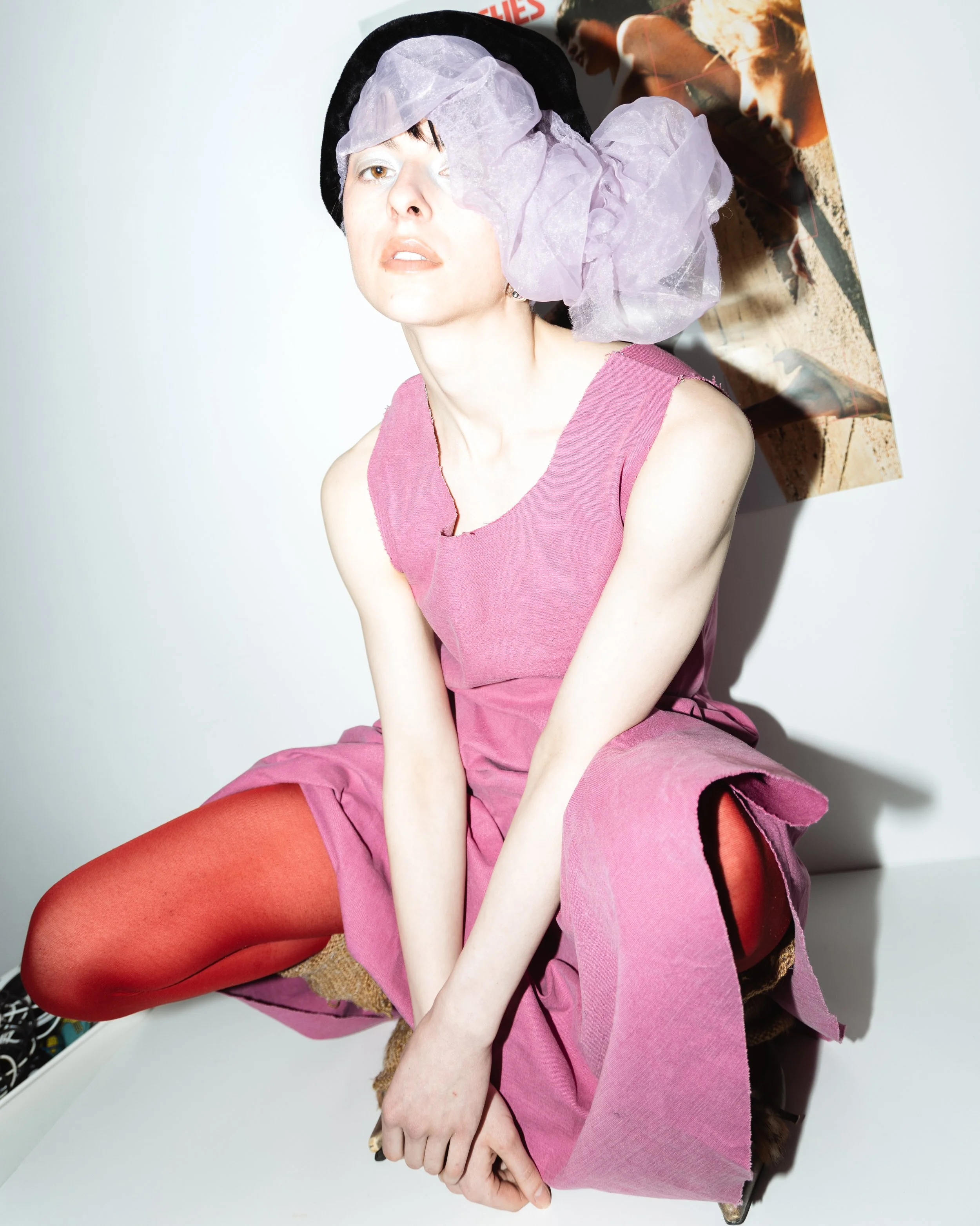Porträt einer jungen Frau mit hellerer Hautfarbe, schwarzen kurzen Haaren und einem schwarzen Hut, die in einem pinken Kleid sitzt. Sie trägt orangefarbene Strümpfe und sitzt auf einem Stuhl vor einem weiße Wand. Im Hintergrund ist eine Zeitschrift m