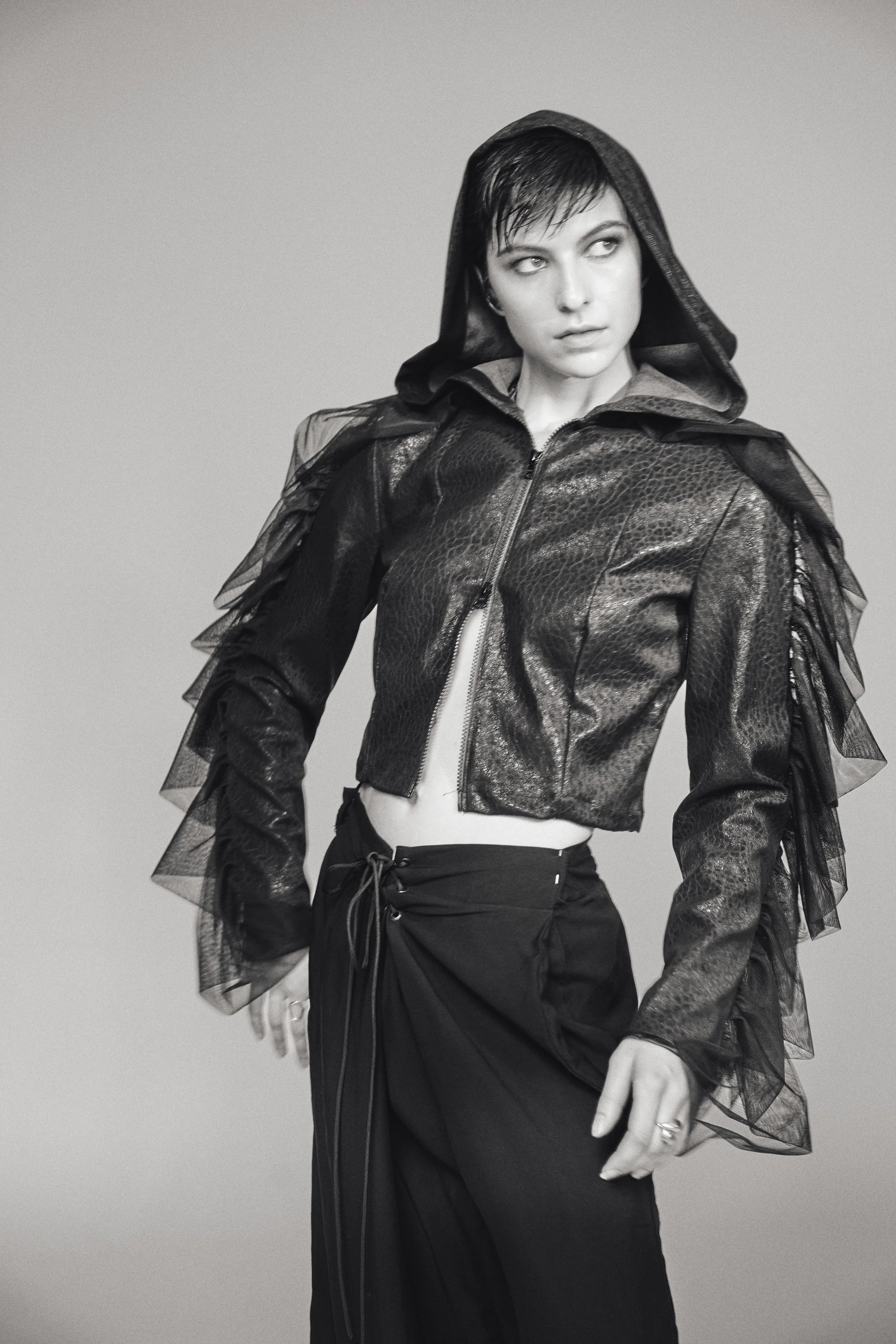 Schwarz-weiß-Foto einer Frau mit kurzen dunklen Haaren, die eine schwarze Jacke mit Tüll-Ärmeln trägt und in einer Pose schaut.