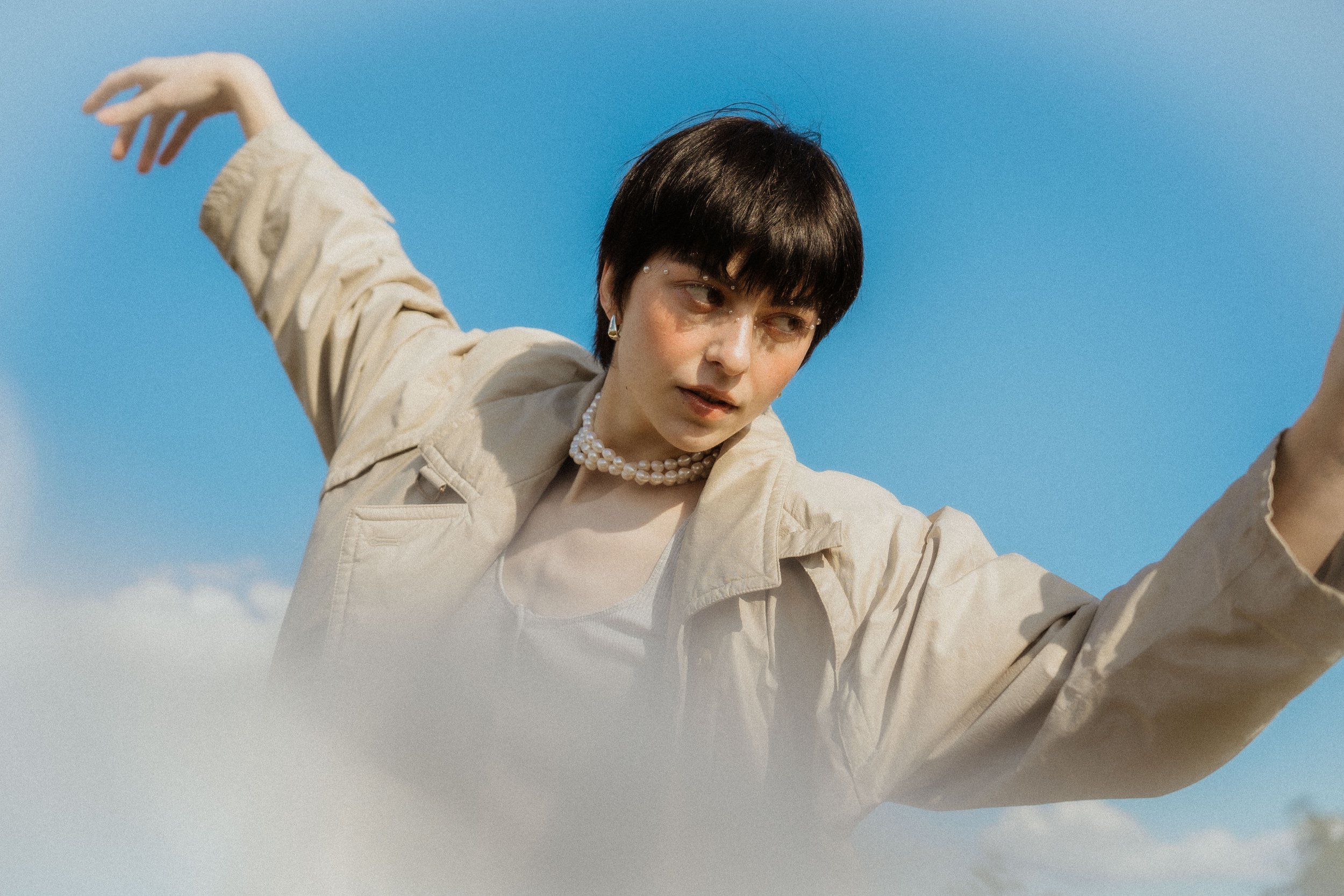 Junge Frau mit dunklem Kurzhaarschnitt, Perlenkette, beige Jacke, vor blauer Himmel, macht eine Pose mit ausgestrecktem Arm.
