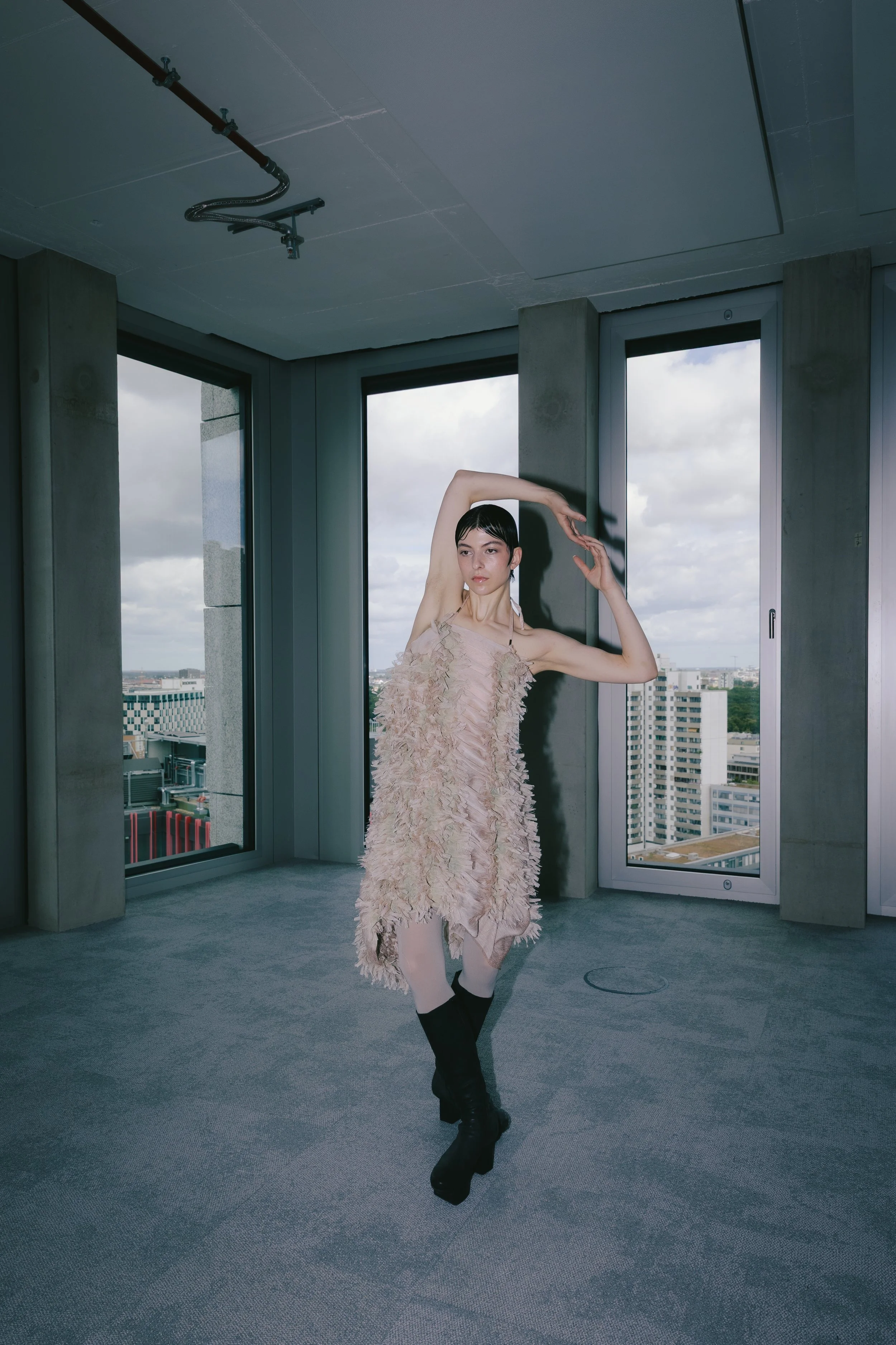 Eine junge Frau tanzt in einem modernen Raum mit großen Fenstern, die eine städtische Skyline zeigen, trägt ein beige, federartige Kleidung und schwarze Stiefel.
