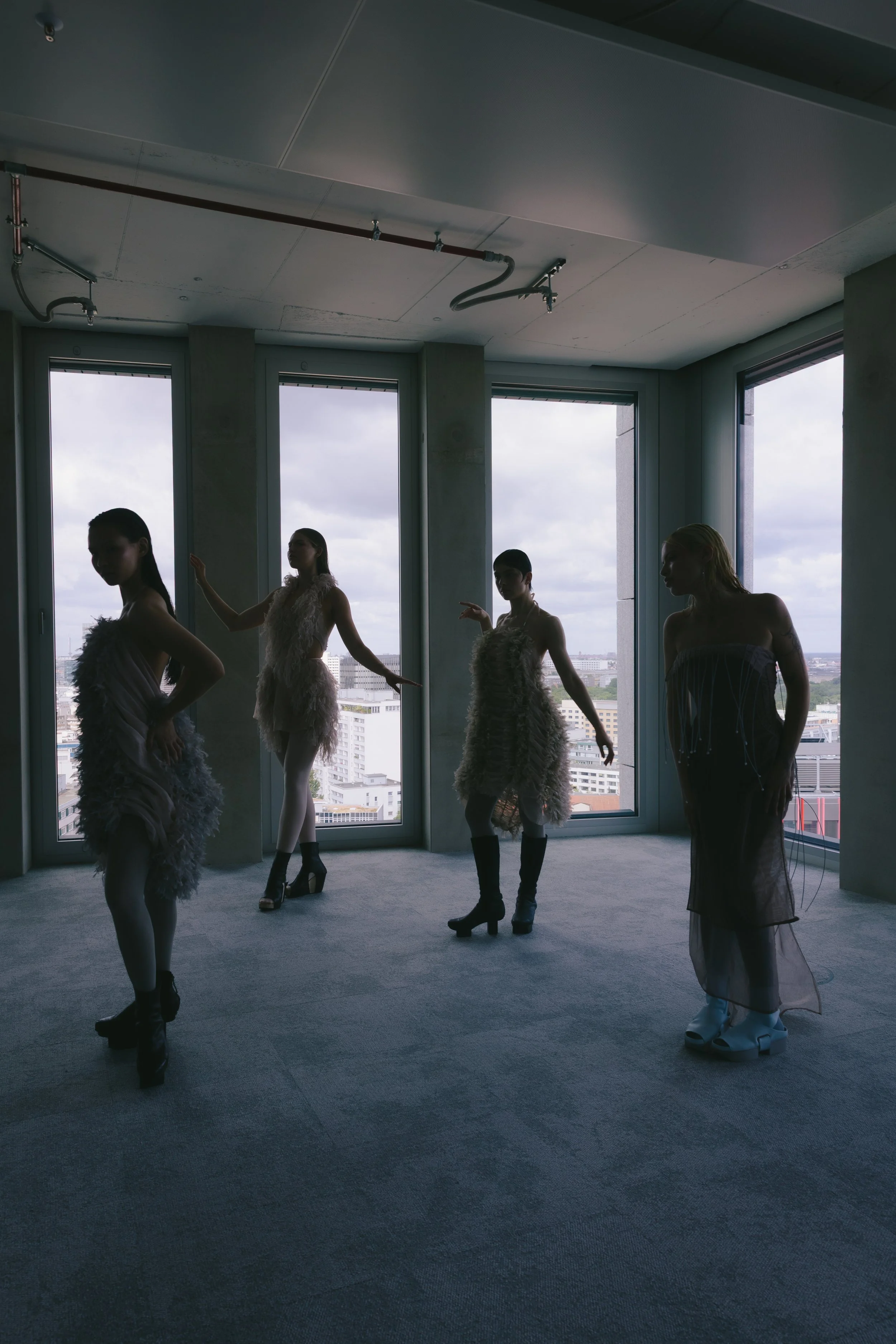 Fünf Frauen posieren in einem modernen, lichtdurchfluteten Innenraum vor großen Fenstern mit Stadtansicht. Sie tragen ausgefallene, elegante Kleider und Schuhmode. Ihre Silhouetten sind dunkel gegen das Tageslicht, das von draußen scheint.