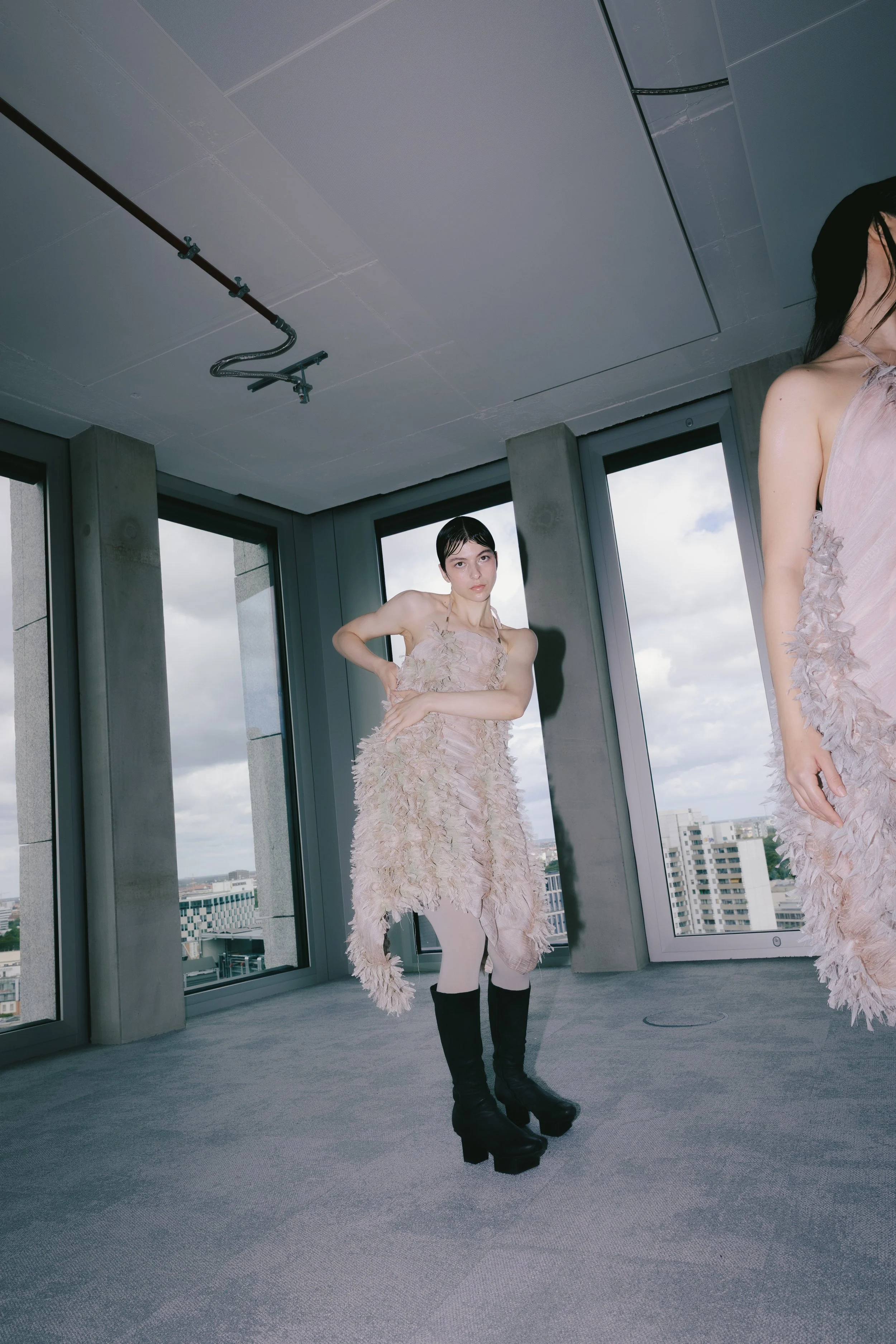 Zwei Frauen in eleganten, nude-farbenen Kleidern mit Federn, die in einem modernen, hoch gelegenen Gebäude mit großen Fensterflächen stehen, umgeben von einer städtischen Skyline.
