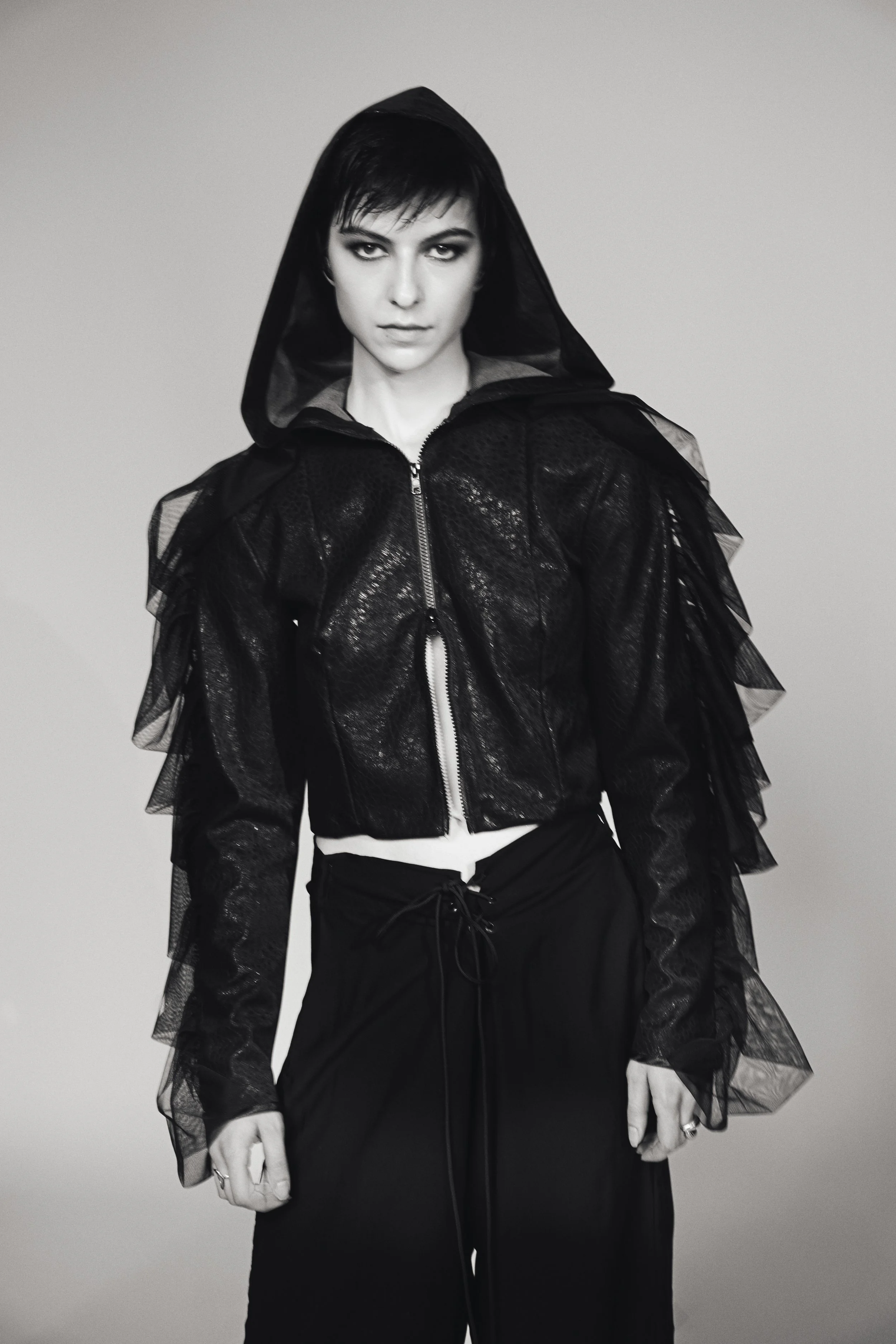 Schwarz-weiß-Foto einer jungen Frau mit kurzen dunklen Haaren, die schwarzen, auffälligen Augen. Sie trägt eine schwarze Jacke mit Tüll-Details an den Schultern und Armen, sowie schwarze Hosen. Der Hintergrund ist schlicht grau.