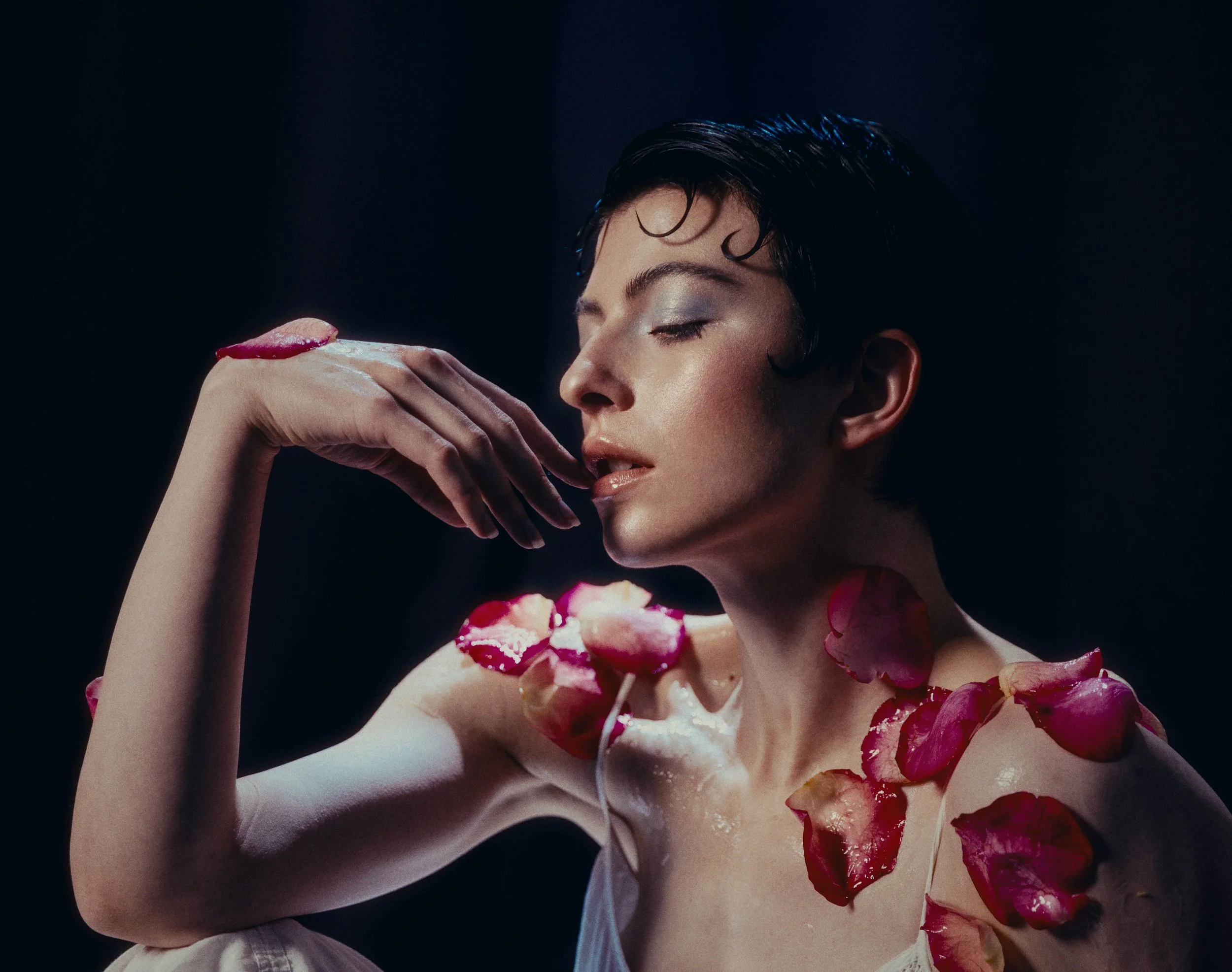 Frau mit nassem schwarzen Haar und glänzendem Make-up, die geschlossen die Augen hält, während sie ein Dessert mit rosa Blüten und weißer Creme isst, vor einem dunklen Hintergrund.