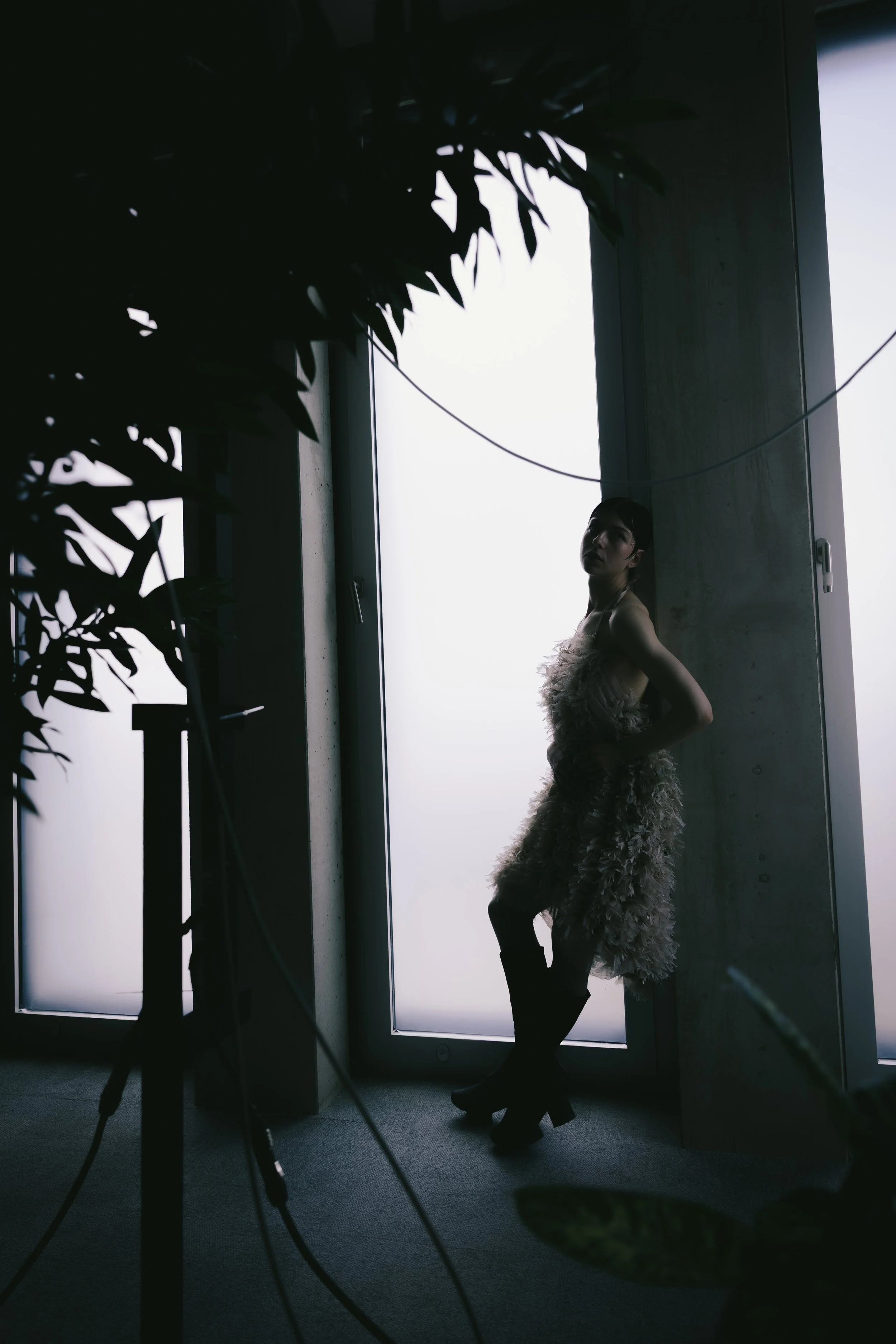Eine Frau in einem auffälligen Kleid posiert vor einem großen Fenster, im Inneren eines Raumes mit dunkler Atmosphäre, umgeben von Kamerastativen und Ausrüstung.