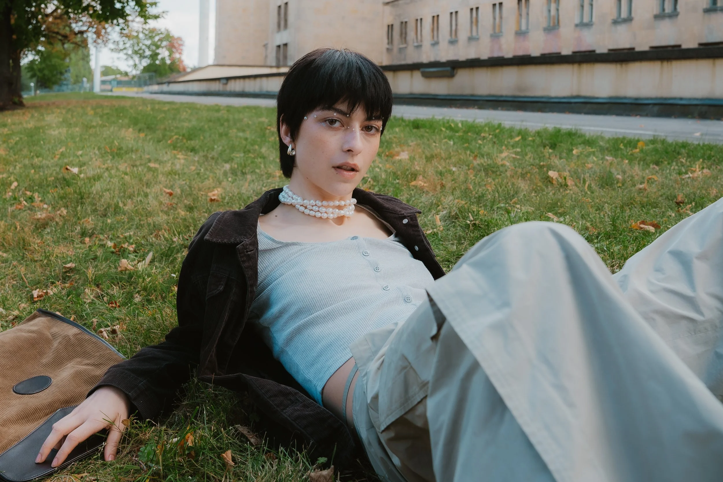 Junge Frau mit dunklem Kurzhaarschnitt, Perl necklace und Piercings auf der Wange, sitzt auf dem Gras vor einem Gebäude mit mehreren Fenstern.