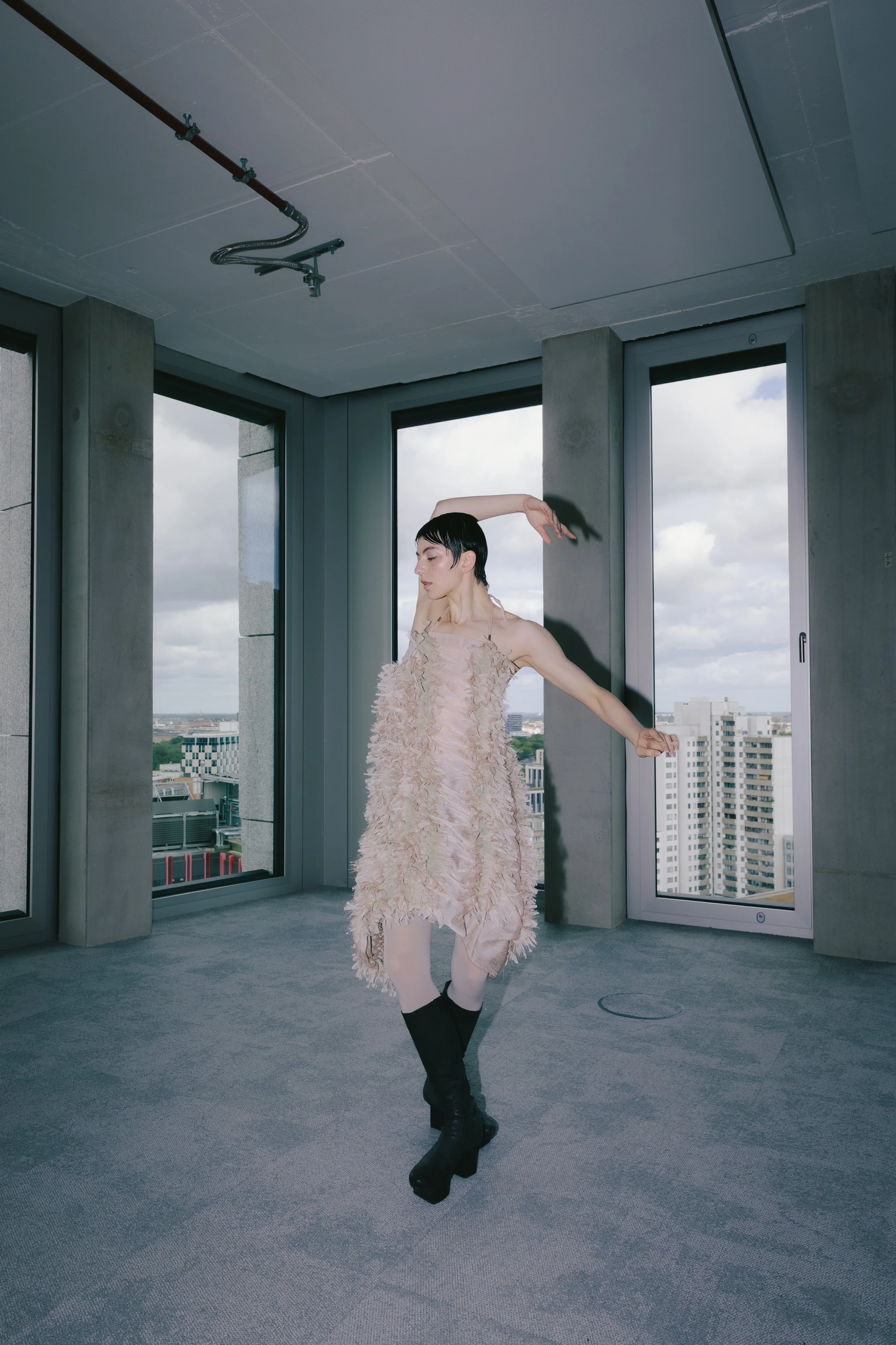 Frau in einem floralen Kleid tanzt in einem modernen, großen Raum mit großen Fenstern und Blick auf die Stadt, trägt schwarze Stiefel.