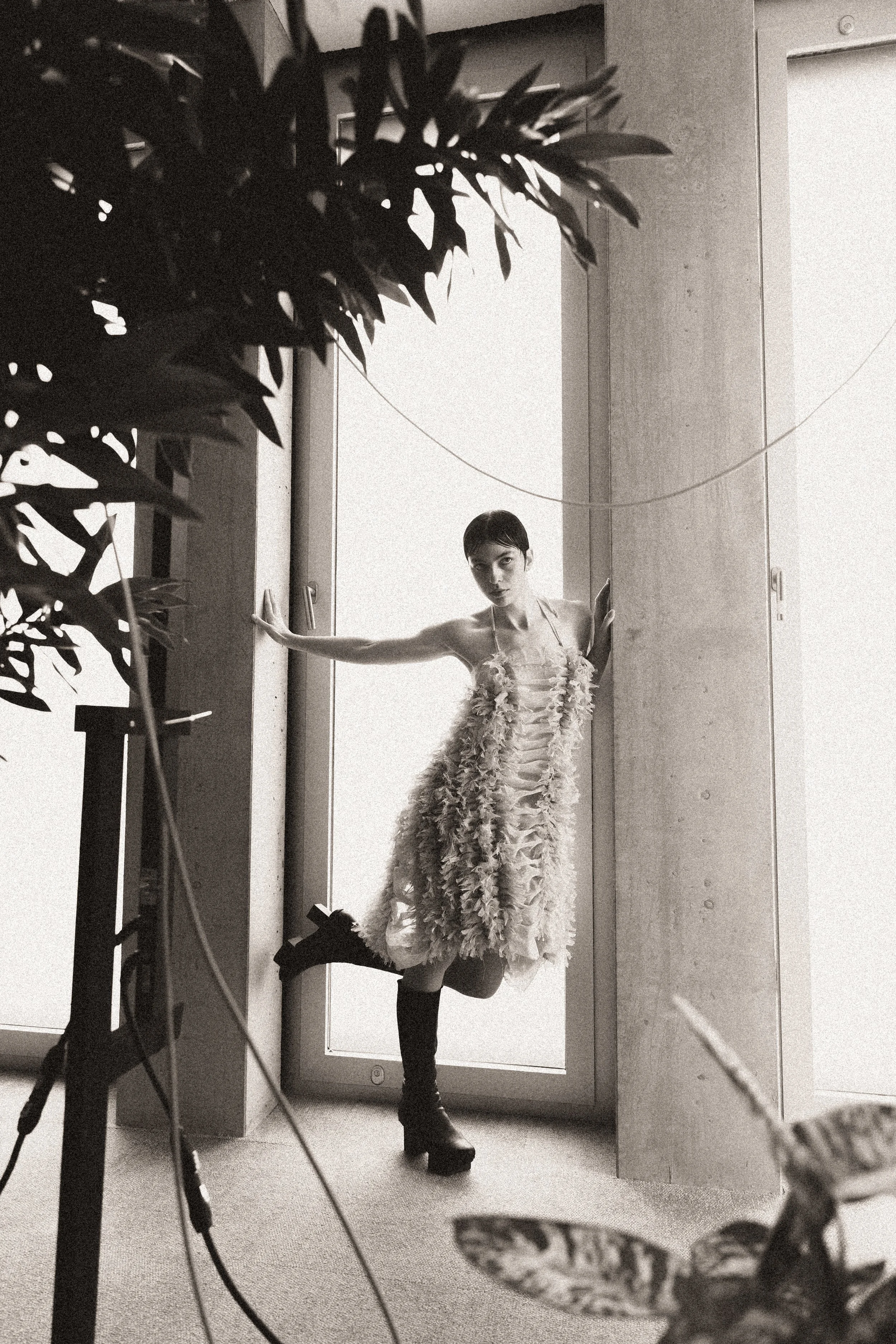 Junge Frau in einem eleganten, verzierter Kleid steht an einem Fenster, während sie Fotoshootings erlebt. Die Szene ist schwarz-weiß und zeigt das Model in einem stilvollen, modischen Outfit.