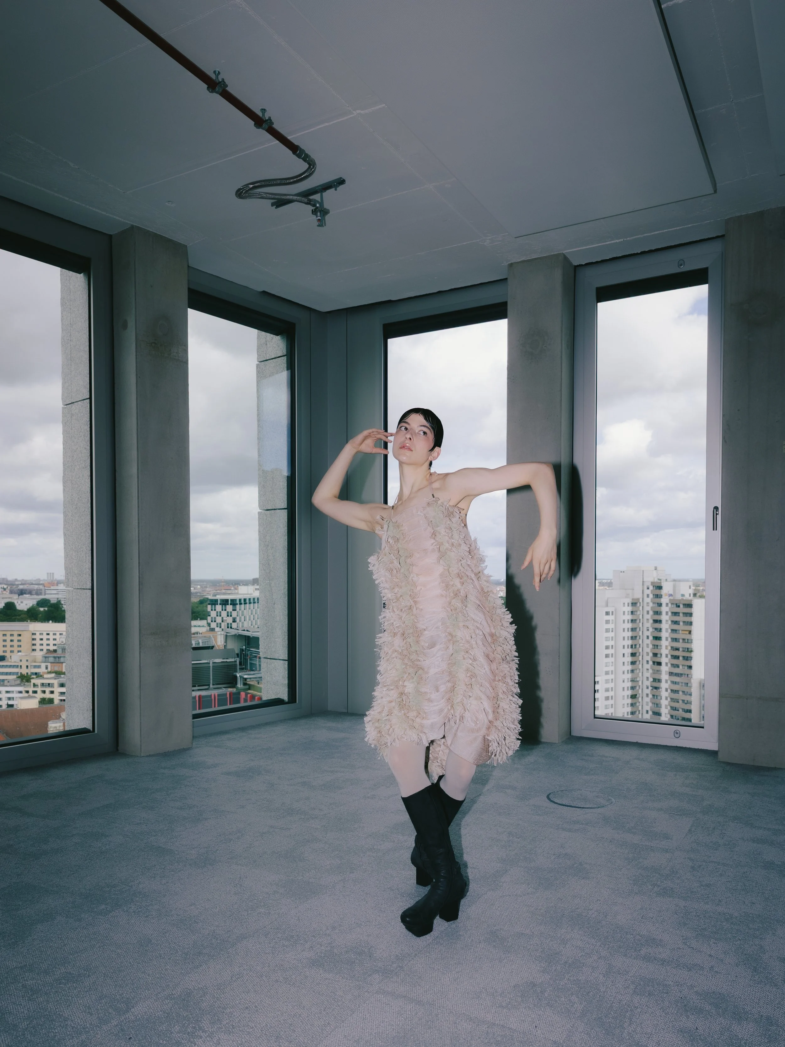 Junge Frau tanzt in einem modernen, hohen Gebäude mit großen Fenstern und Stadtblick, trägt ein elegantes, pfirsichfarbenes Kleid mit Federn und schwarze Stiefel.
