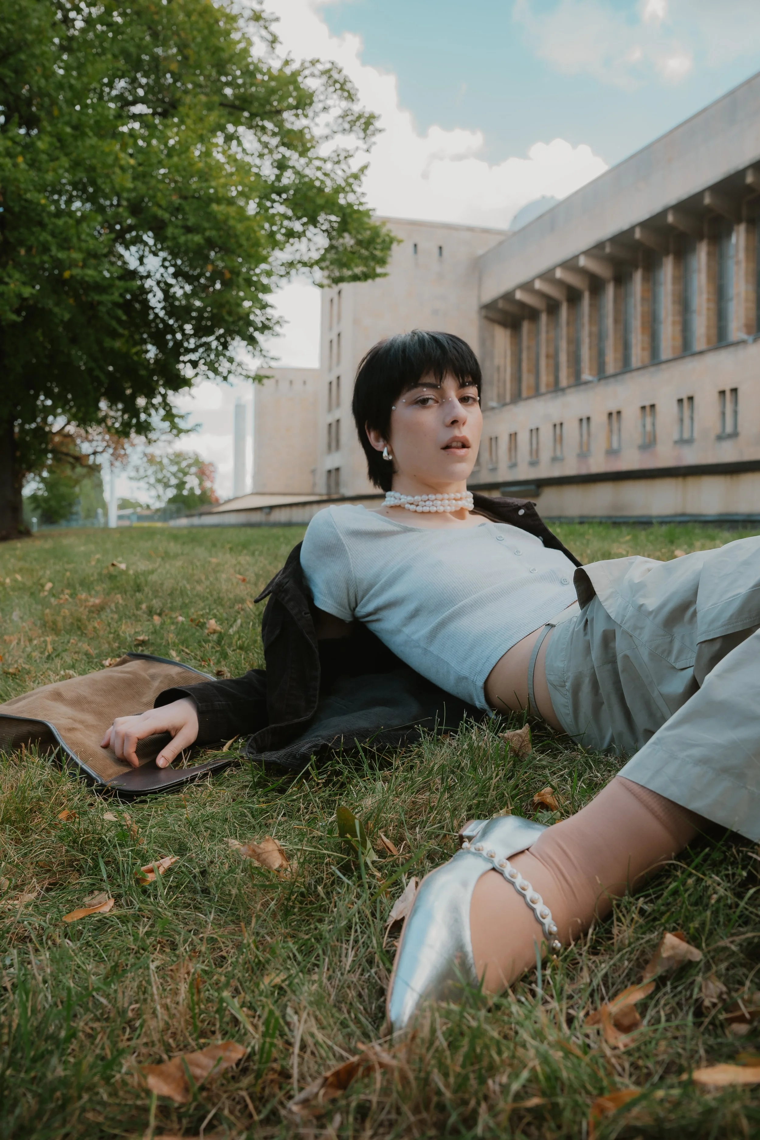 Junge Frau liegt entspannt auf dem Rasen vor einem modernen Gebäude, trägt eine Perlenkette und einen weißen T-Shirt, im Hintergrund Bäume und Himmel.