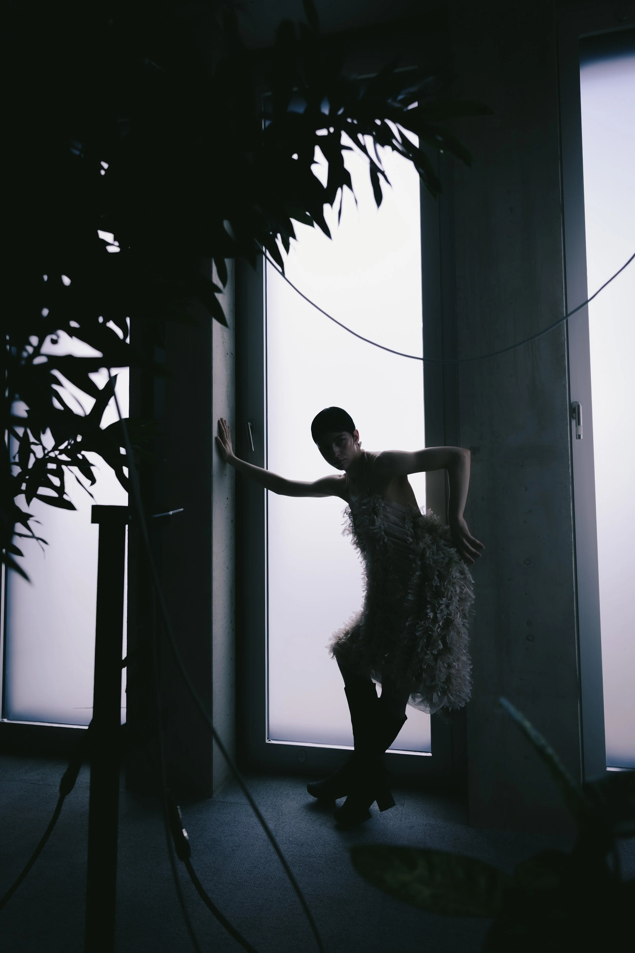 Silhouette eines Models in einem dunklen Raum vor einem hellen Fenster, trägt ein auffälliges, texturiertes Kleid und steht in Pose, während Fotografen und Kameraausrüstung im Vordergrund sind.