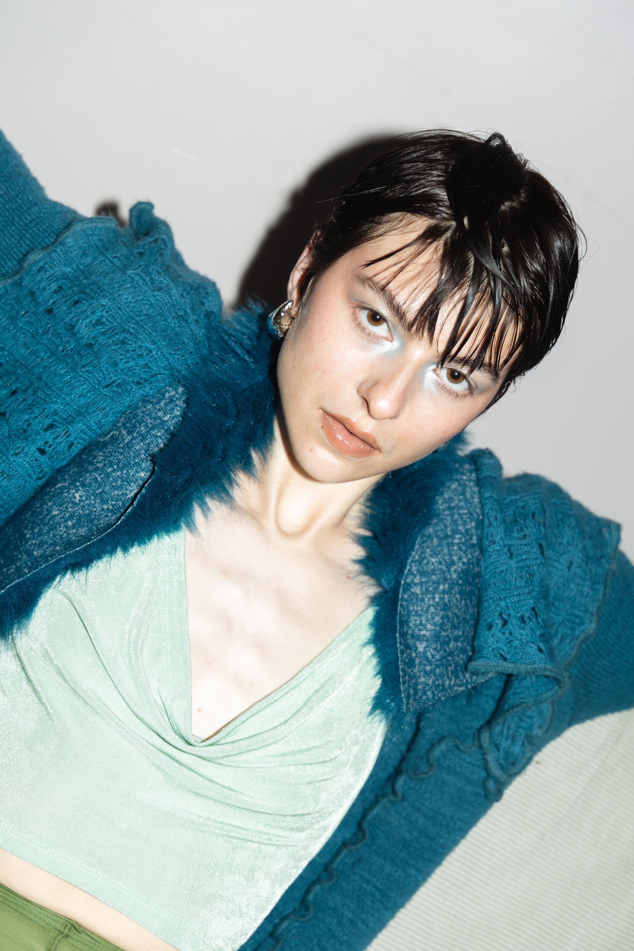 Ein Porträt einer jungen Frau mit dunklen, nassen Haaren, blauen Augen und einem blonden Hautton, die in einem grauen Raum posiert, trägt ein grünes Top und einen blauen Strickpullover.