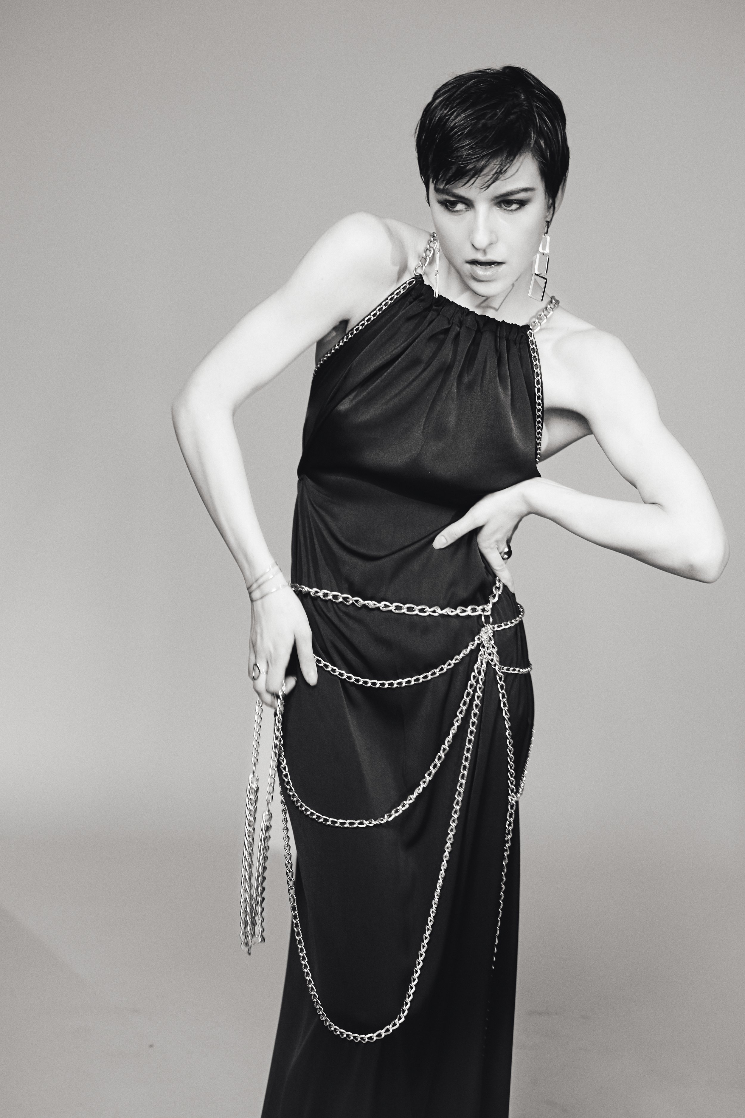 Schwarz-Weiß-Foto einer jungen Frau mit kurzer dunkler Frisur, die ein elegantes schwarzes Kleid trägt, verziert mit mehreren silbernen Ketten und Ohrringen, und eine nachdenkliche Pose einnimmt.