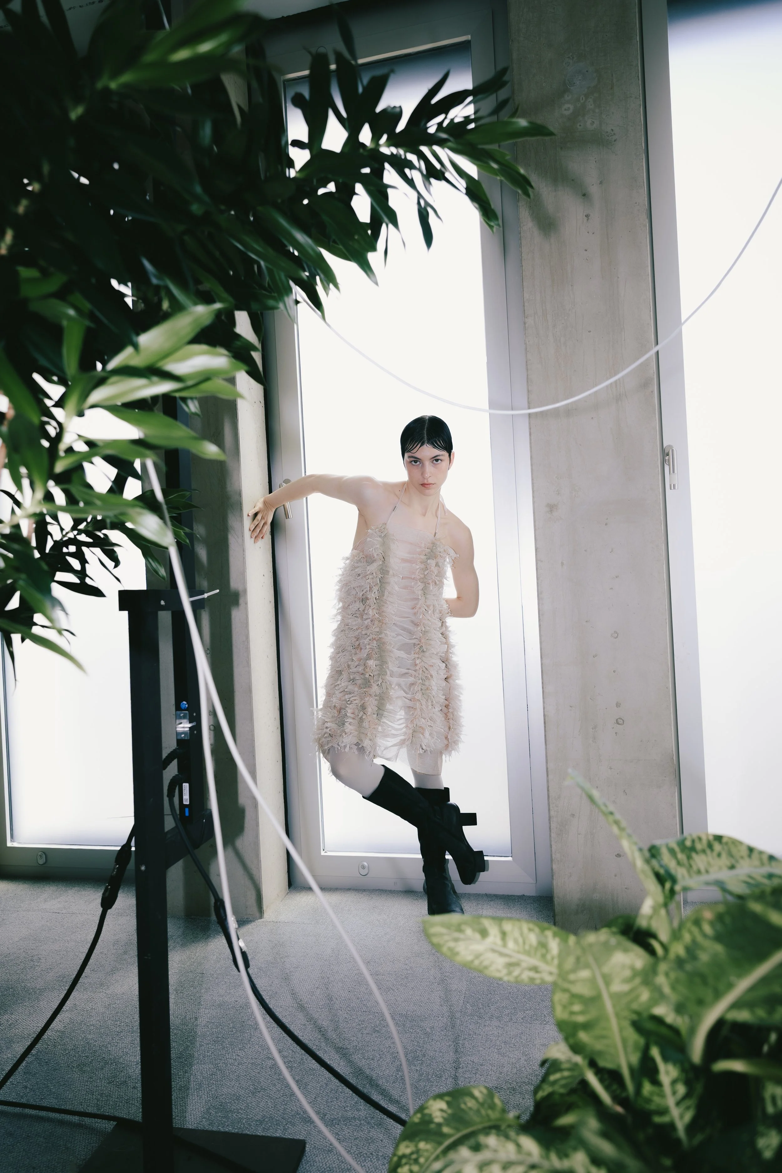 Junge Frau mit schwarzen Haaren und modebewusstem Outfit steht in einem modernen Innenraum vor einer Fenstertür, umgeben von Pflanzen, bei einem Fotoshooting.