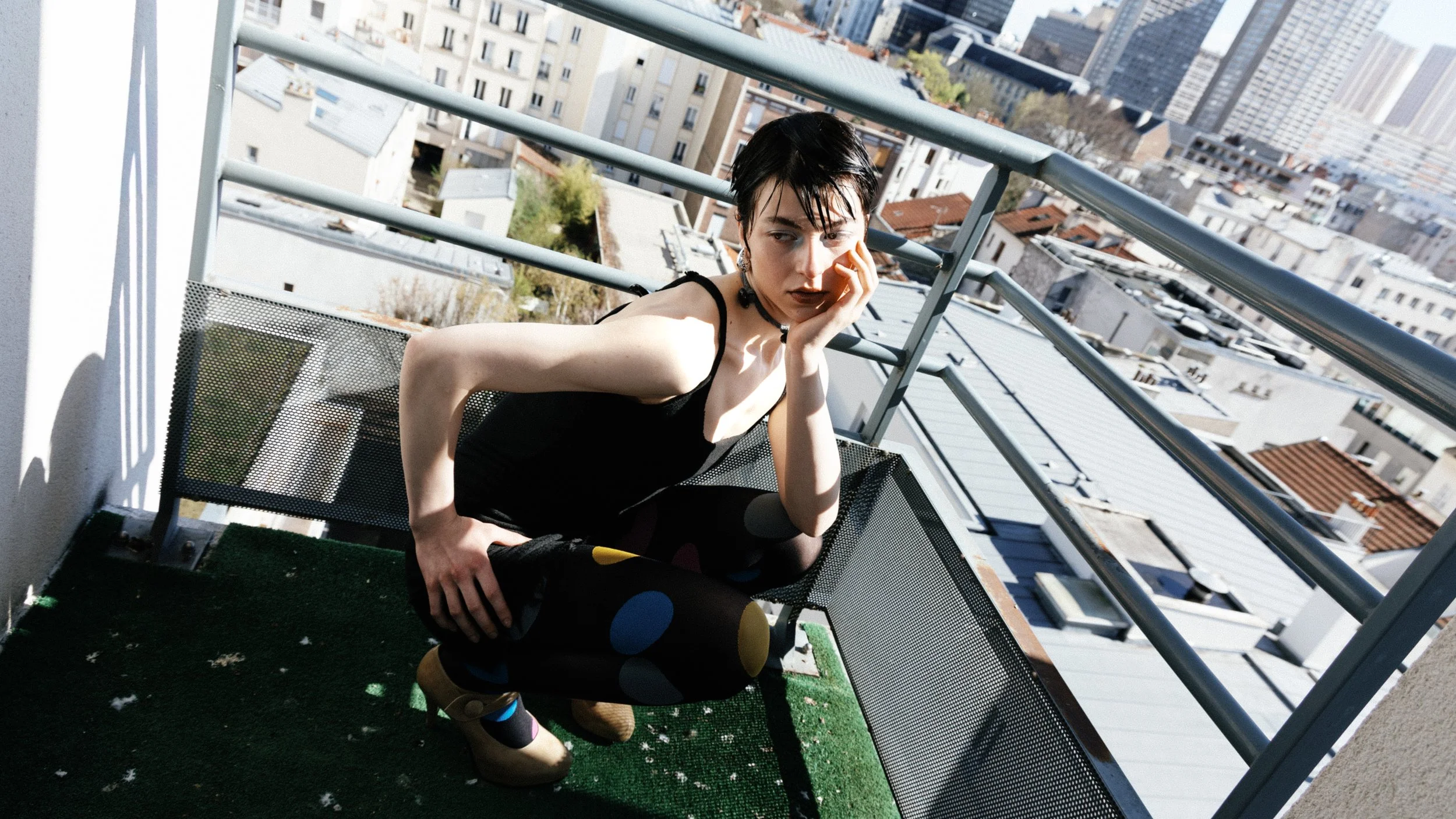 Junge Frau mit schwarzen Haaren in einem schwarzen Kleid sitzt auf einem Balkon und schaut nach unten, mit städtischer Skyline im Hintergrund.