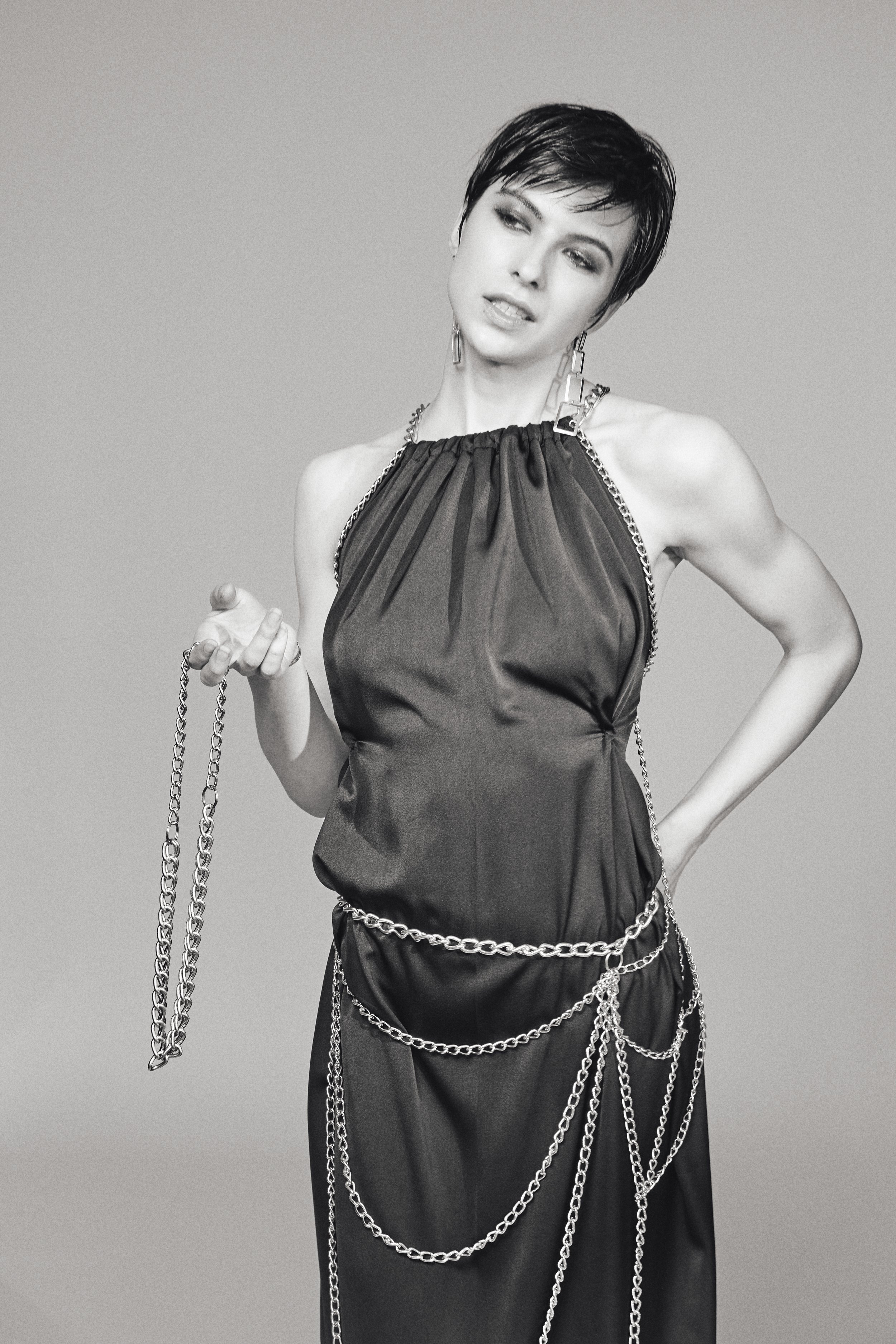 Schwarz-weiße Fotografie einer Frau mit kurzen schwarzen Haaren, die eine dunkle geraffte Kleid trägt und mit mehreren silbernen Ketten geschmückt ist. Sie blickt seitlich, mit einem leichten Lächeln, vor grauem Hintergrund.