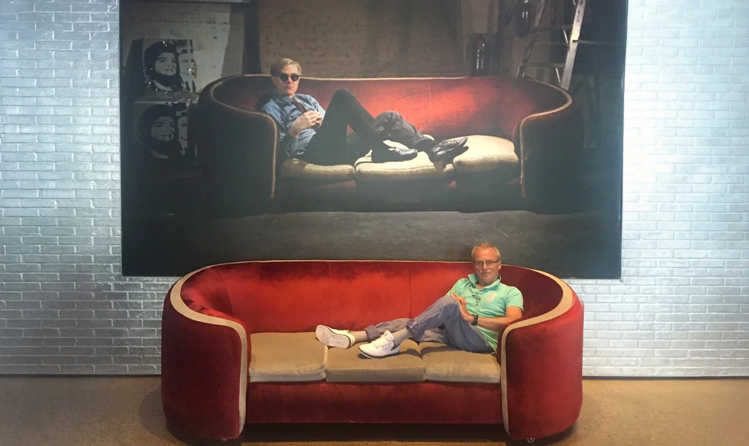 Ein Mann sitzt auf einem roten, halbkreisförmigen Sofa vor einer großen Wand mit einem Gemälde. Auf dem Gemälde ist eine Person mit Sonnenbrille auf einem Sofa zu sehen.