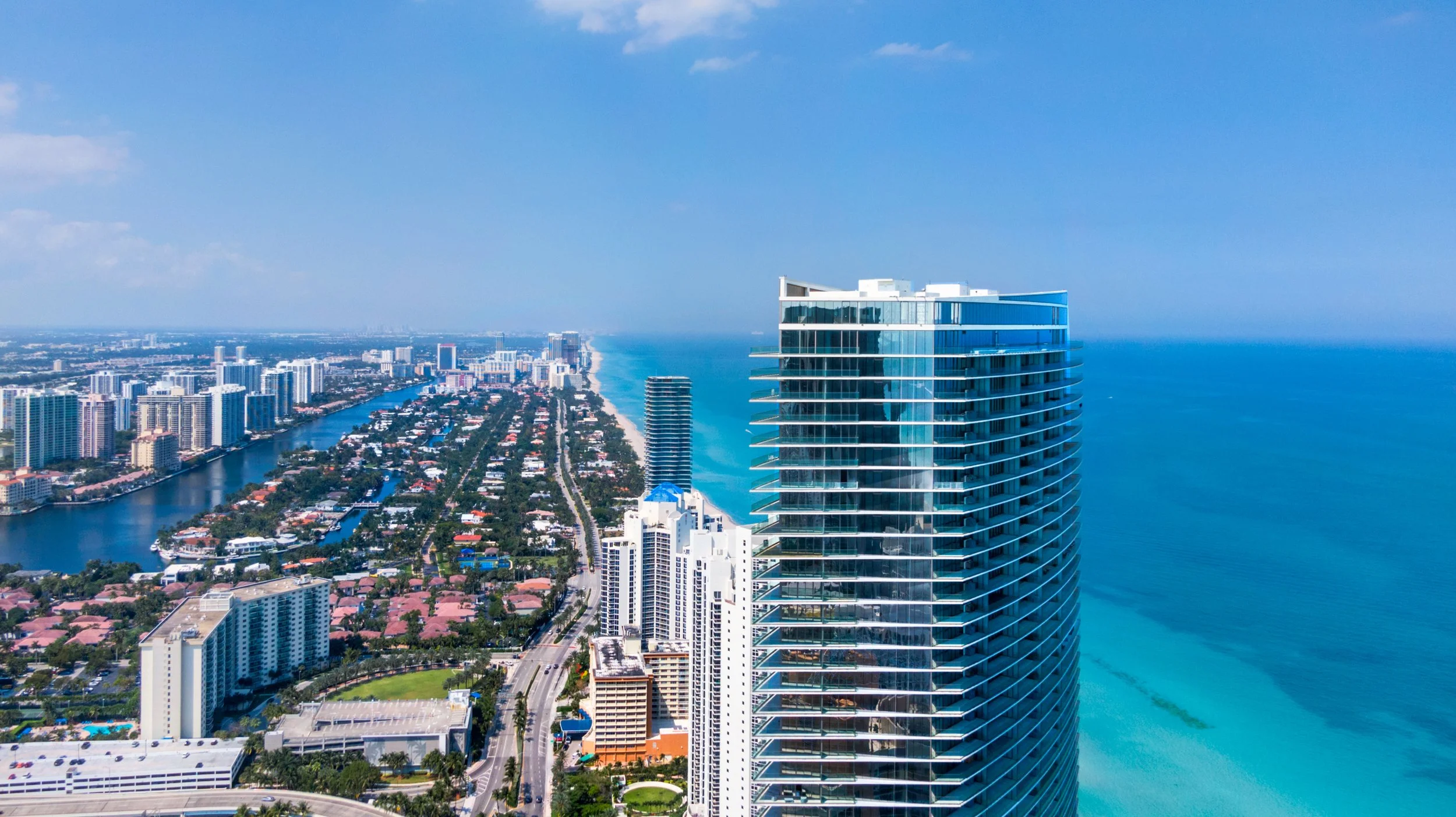 A high-rise modern building with glass balconies overlooking a coastal city with many skyscrapers, waterways, and a sandy beach along the ocean.