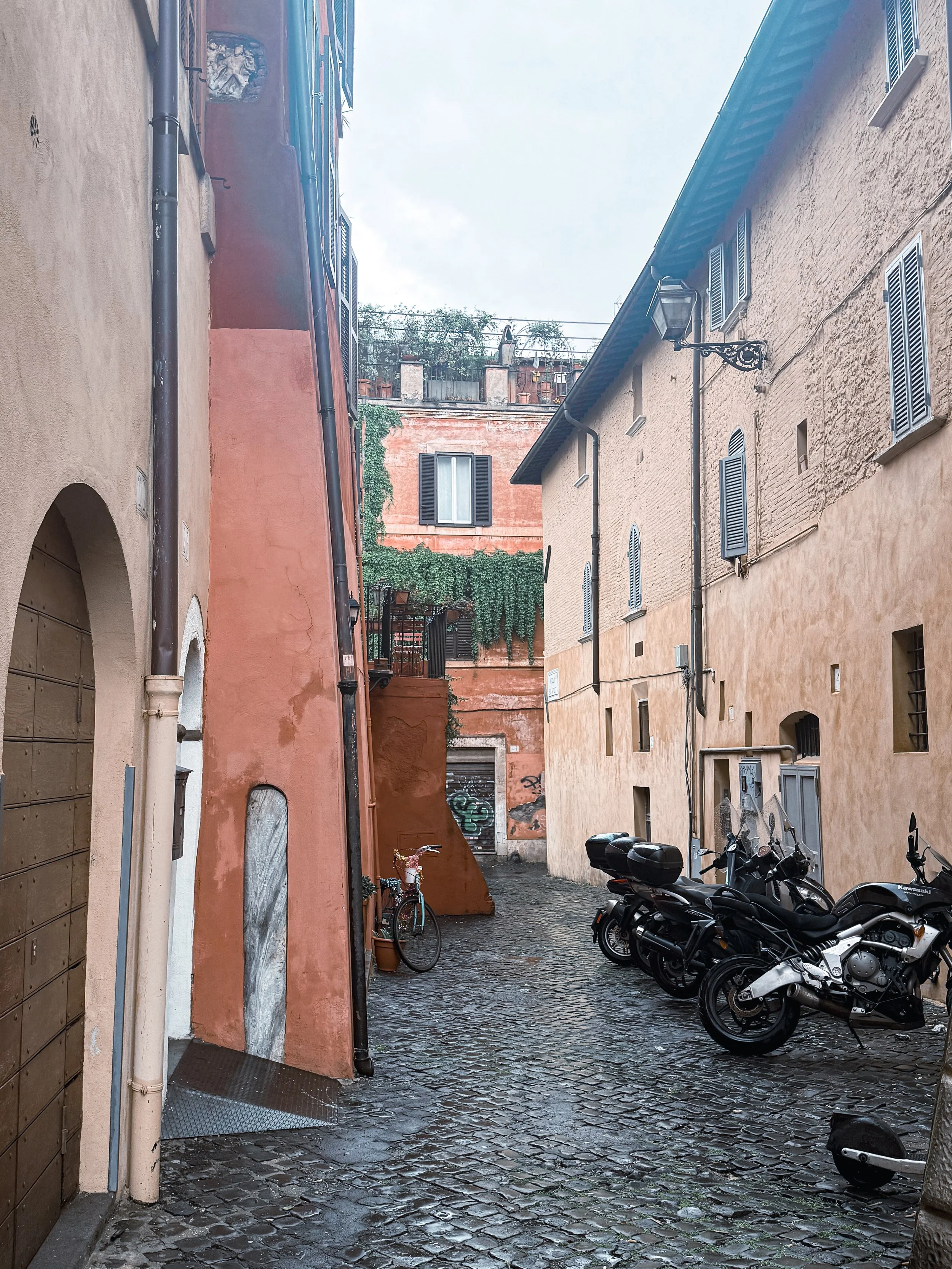 A narrow cobblestone alleyway with pastel-colored buildings on both sides, parked motorcycles, and a bicycle near a orange wall with greenery and graffiti.