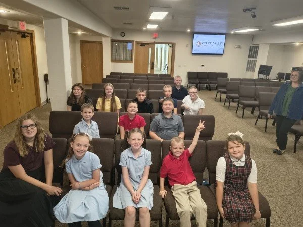 A group of children and adults sitting in a church or community room, some smiling and making peace signs, with rows of empty chairs and a screen displaying the words "Pine Valley" in the background.