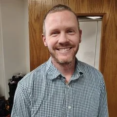 A smiling man with short hair and a beard, wearing a blue checkered shirt, standing indoors against a wooden door and beige wall.