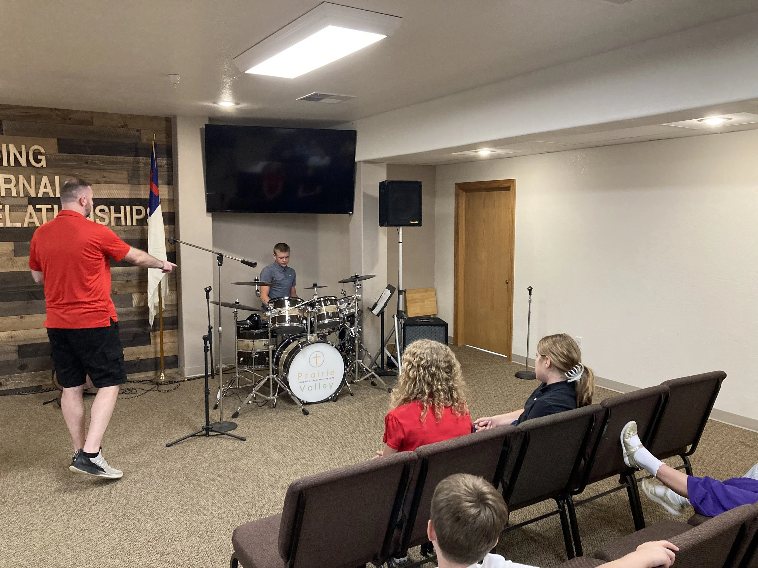 Group of children and adults seated in a room with musical instruments, including drums, during a church or community event.