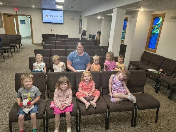A group of nine children and one adult girl sitting and standing in a church or community room with chairs arranged for a gathering, a television screen on the wall displaying 'PRAIRIE VALLEY,' and colorful stained glass windows.