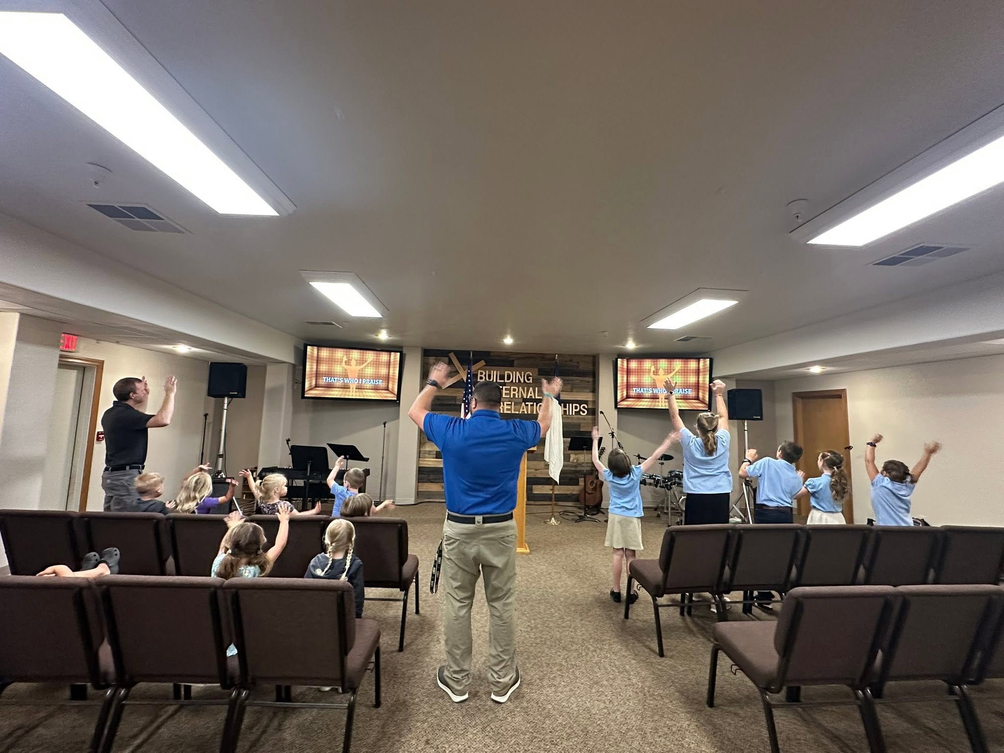 People gathered in a church auditorium participating in a worship service, with some raising their hands, and a large screen displaying song lyrics.