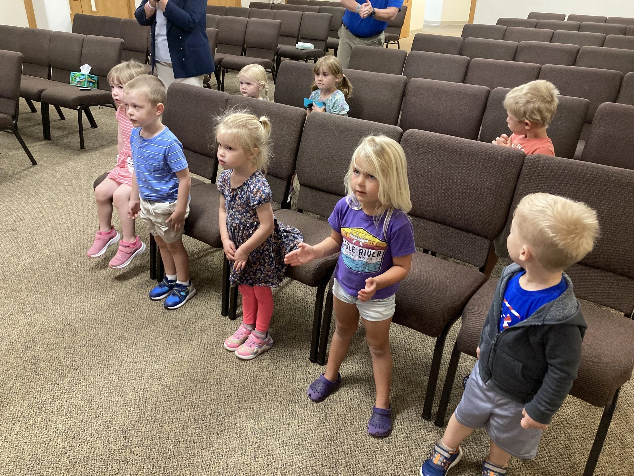 Children standing in a row inside a church or meeting hall with brown chairs, some adults standing in the background.