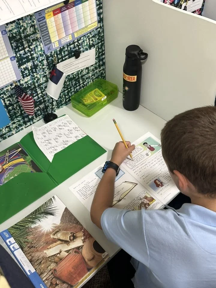 A child at a school desk reading a book with colorful illustrations and writing, surrounded by school supplies including a green folder, a plastic pencil case, a water bottle, and notes on a bulletin board.