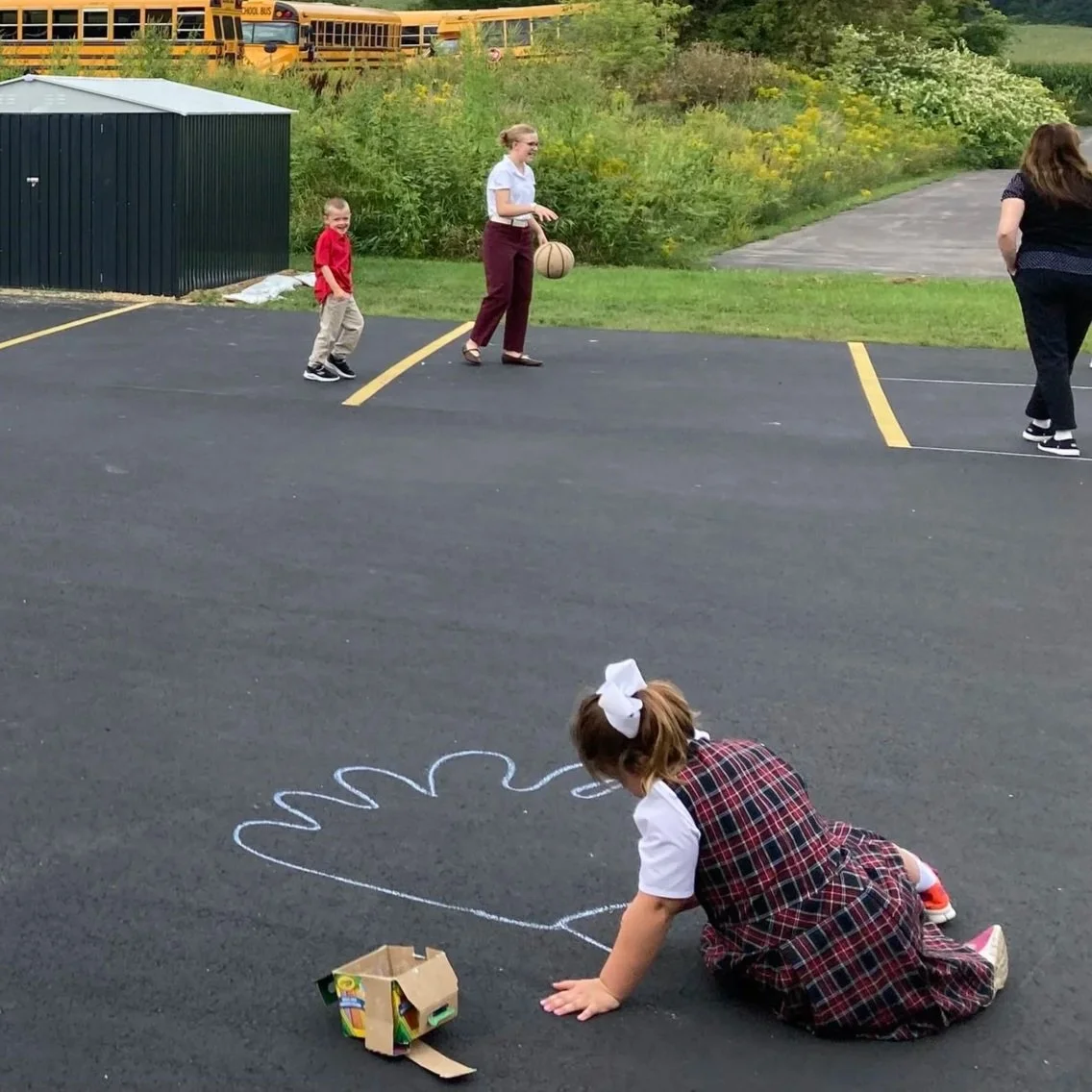 A young girl in a plaid dress is drawing a turkey on the pavement with chalk. Other children and an adult are in the background, with a row of school buses parked behind a grassy area and trees.