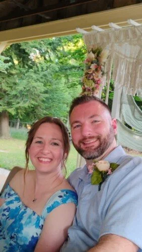 A smiling couple taking a selfie outdoors during the daytime. The woman has long hair and is wearing a blue floral dress. The man has short hair and a beard, wearing a light blue shirt with a boutonnière. Behind them is a decorated backdrop with flowers and a white curtain, with green trees and sunlight visible in the background.