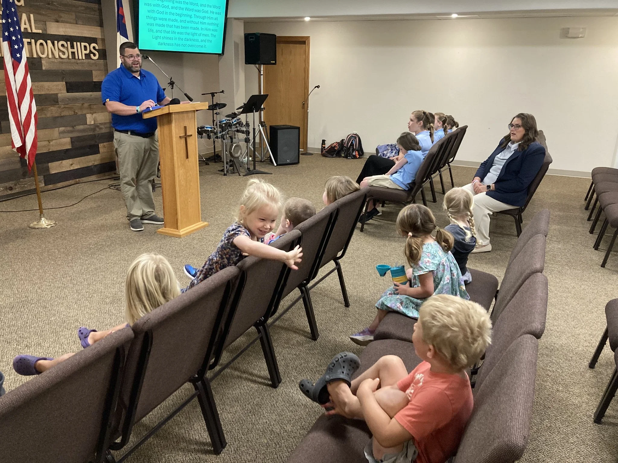 A man in a blue shirt standing at a wooden podium with a cross symbol, speaking to a group of children and a woman in a classroom or church setting. An American flag is on the left, and a large screen with text is in the background. The children are sitting in chairs, some are looking at the speaker, and some are engaging with toys or talking to each other.