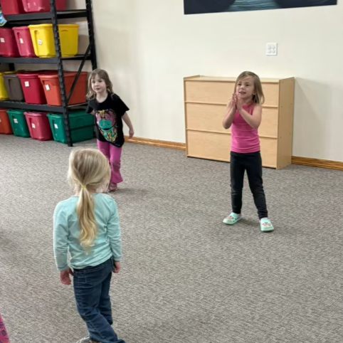 Three young girls are playing indoors, with one girl facing away in the foreground and two girls standing further back, one with hands together and the other with hands near her face, in a room with storage bins and a wooden dresser.