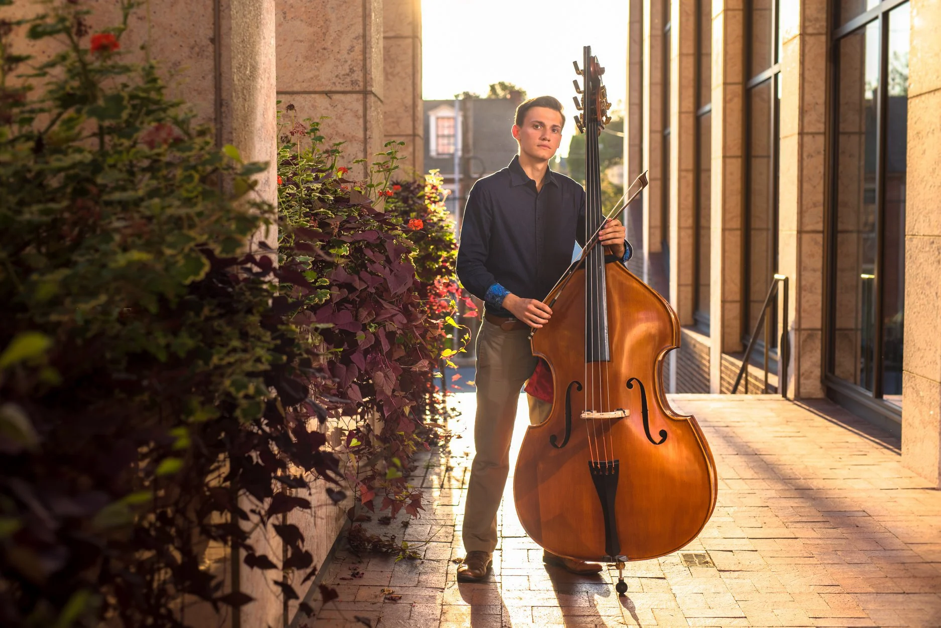 A young man standing outdoors on a brick sidewalk at sunset at the WARE center in Lancaster PA, holding a double bass and a bow, with building windows and a wall with plants beside him.