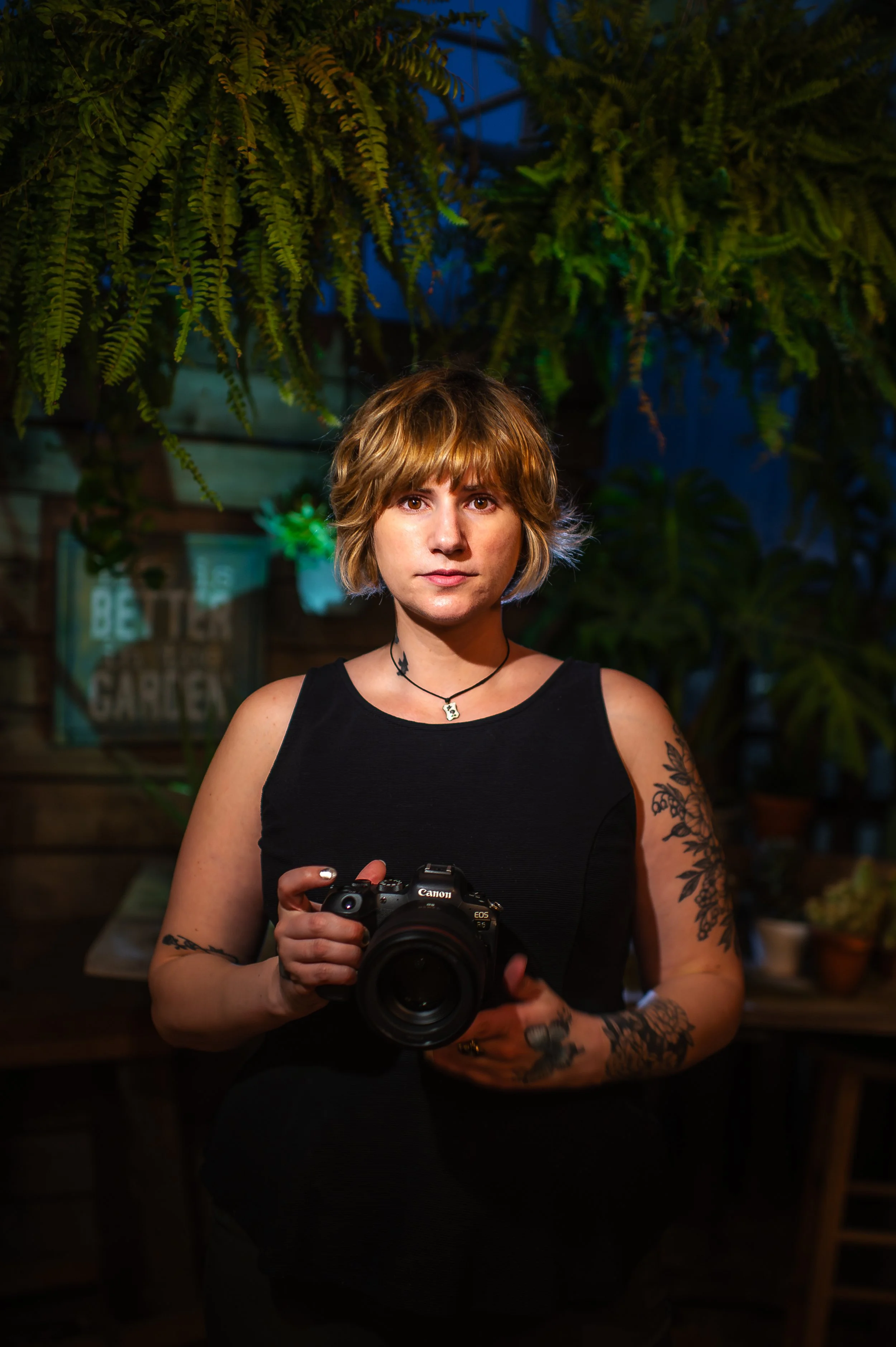 A woman with short blonde hair holding a professional camera in a dimly lit environment with hanging green plants in the background.