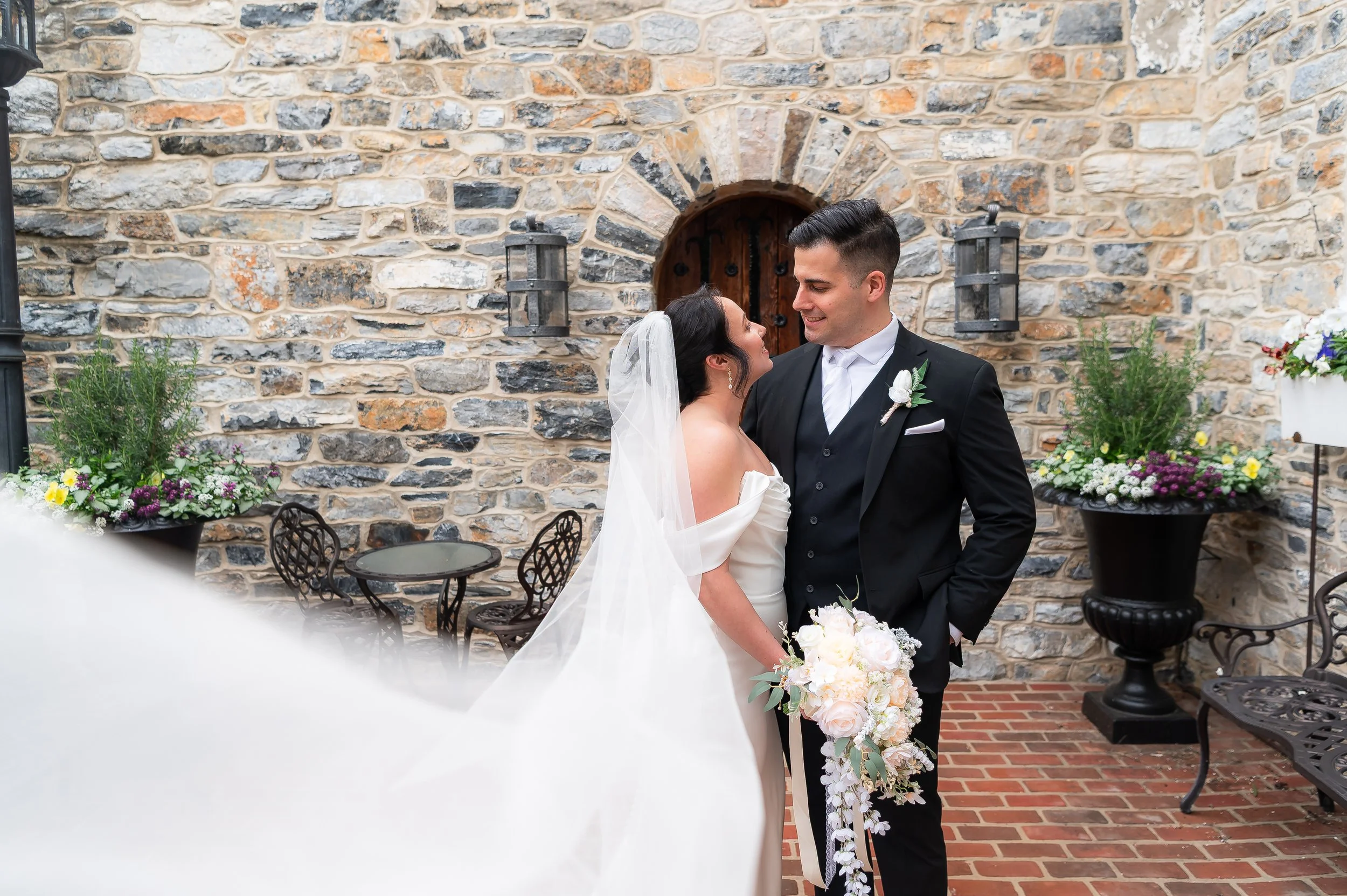 Bride and groom smiling at each other outdoors against a brick wall at Stone Gables Estate's Star Barn, Elizabethtown PA. The bride holds a bouquet of white and blush flowers; the groom wears a black tuxedo with a white boutonniere.