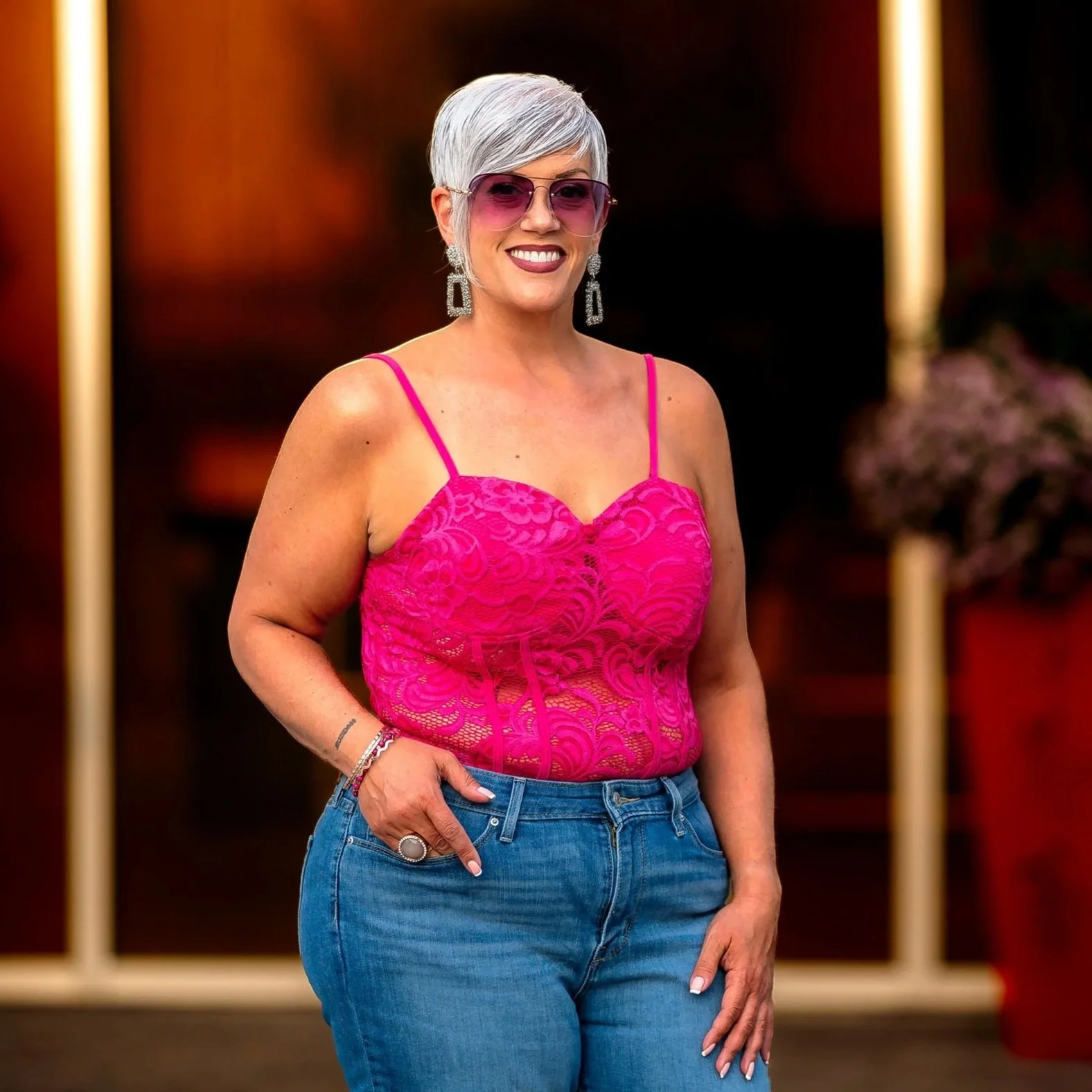 Smiling woman with short silver hair wearing pink sunglasses and a pink lace top, standing indoors at Ewell Plaza in Lancaster, PA.