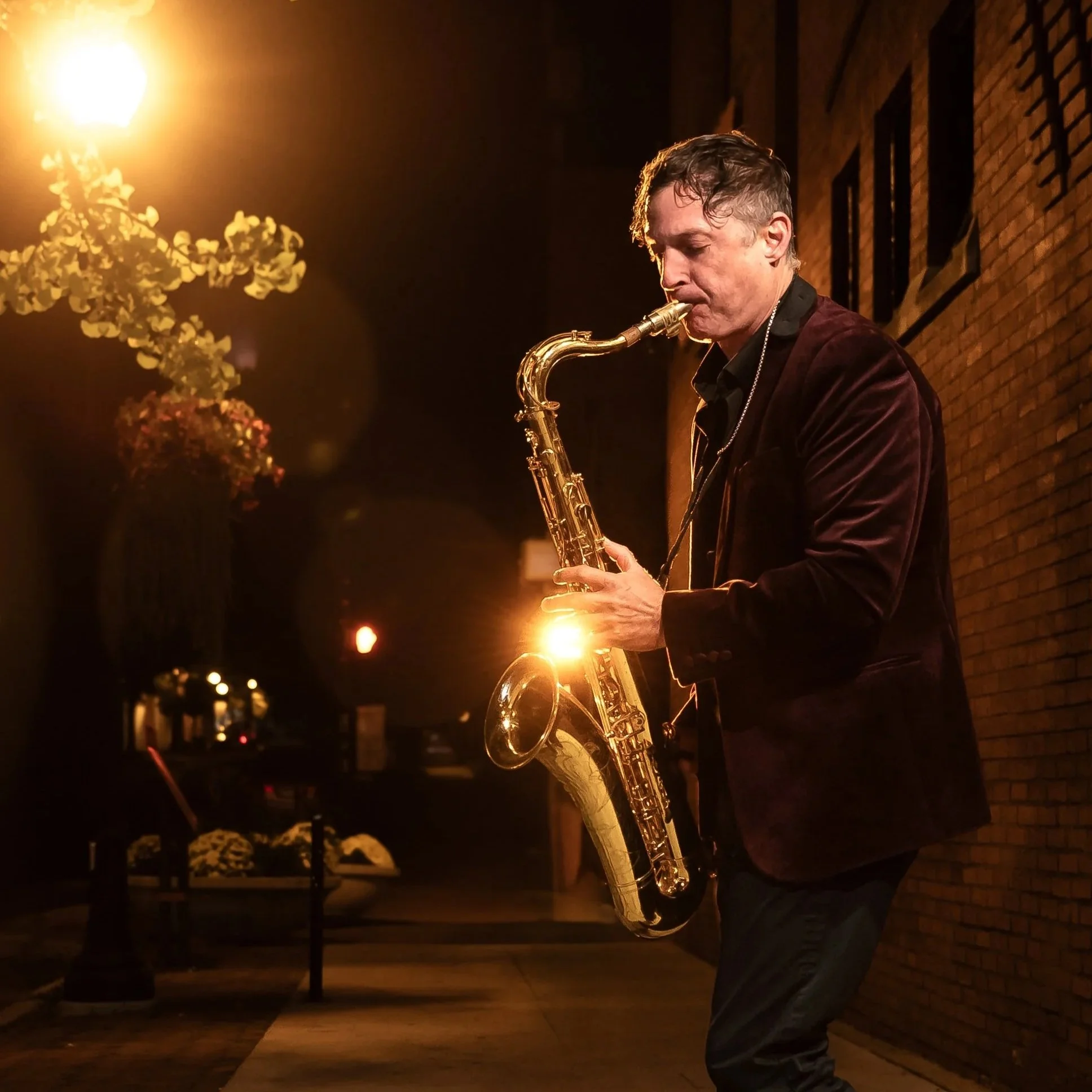 A man is playing a saxophone on a dimly lit street at night, with warm lighting from a nearby streetlamp and a brick wall in the background.